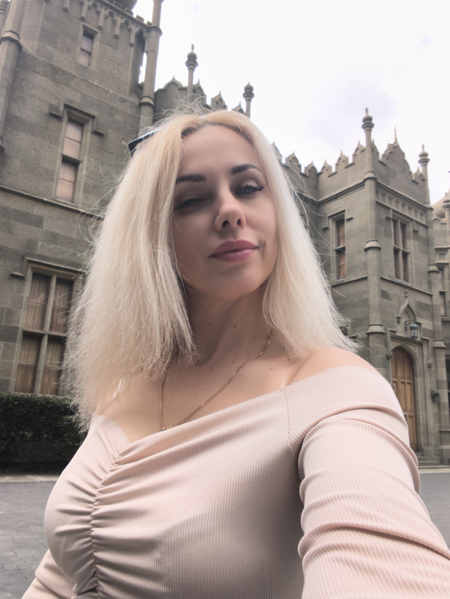 A woman with blonde hair is taking a selfie in front of the Vorontsov Palace in the city of Алупкинский городской совет, Ukraine. She is wearing a light pink off-the-shoulder top and a gold necklace. The palace, a large grey stone building with multiple windows and towers, is behind her. There is some green foliage visible on the left side of the frame. The sky is overcast.