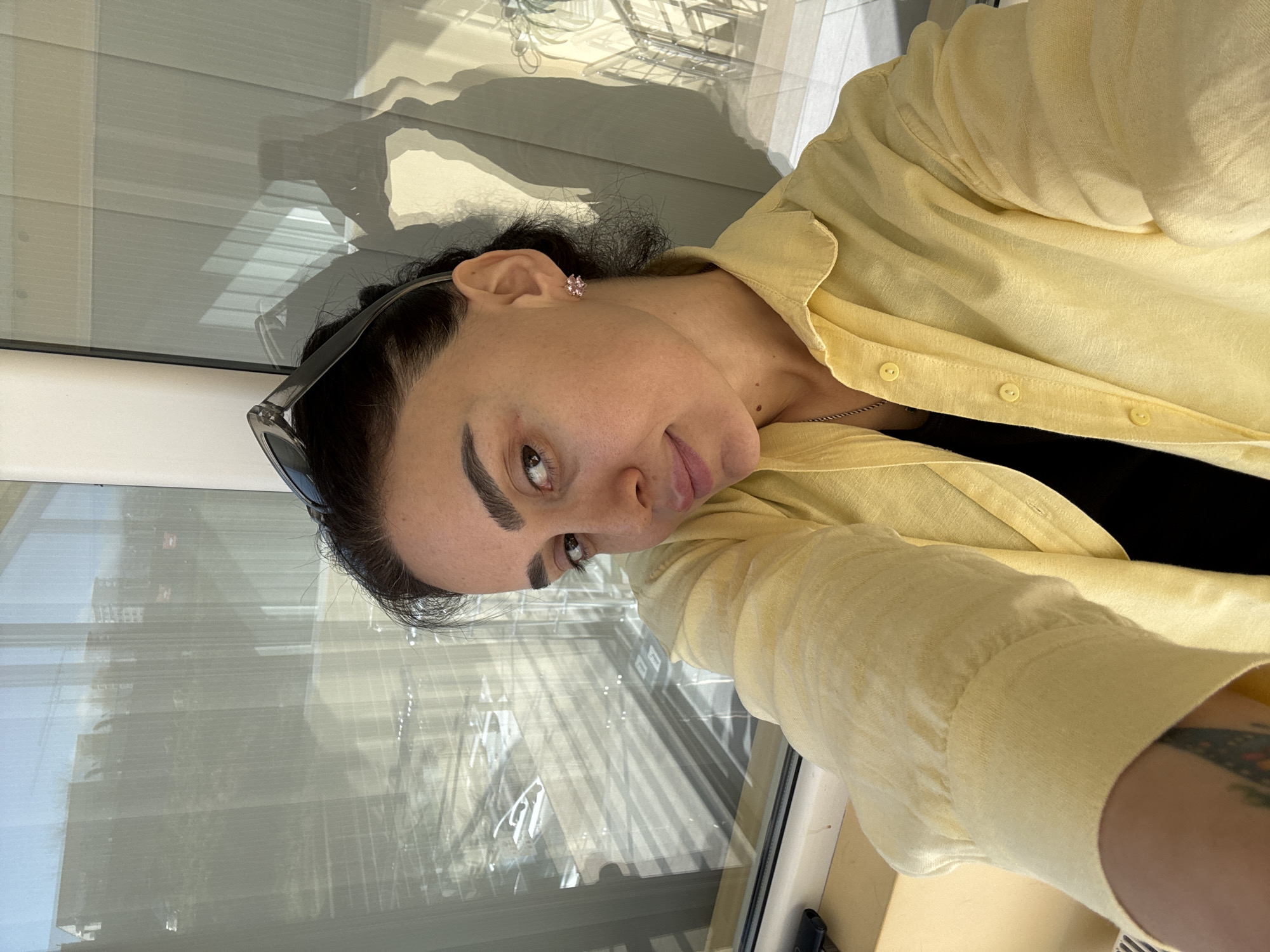 A woman is taking a selfie. She has dark hair, sunglasses resting on her head, and is wearing a light yellow button-down shirt over a black top. She is standing near a window with horizontal blinds. Outside the window, a cityscape is reflected, possibly in Tbilisi, Georgia.