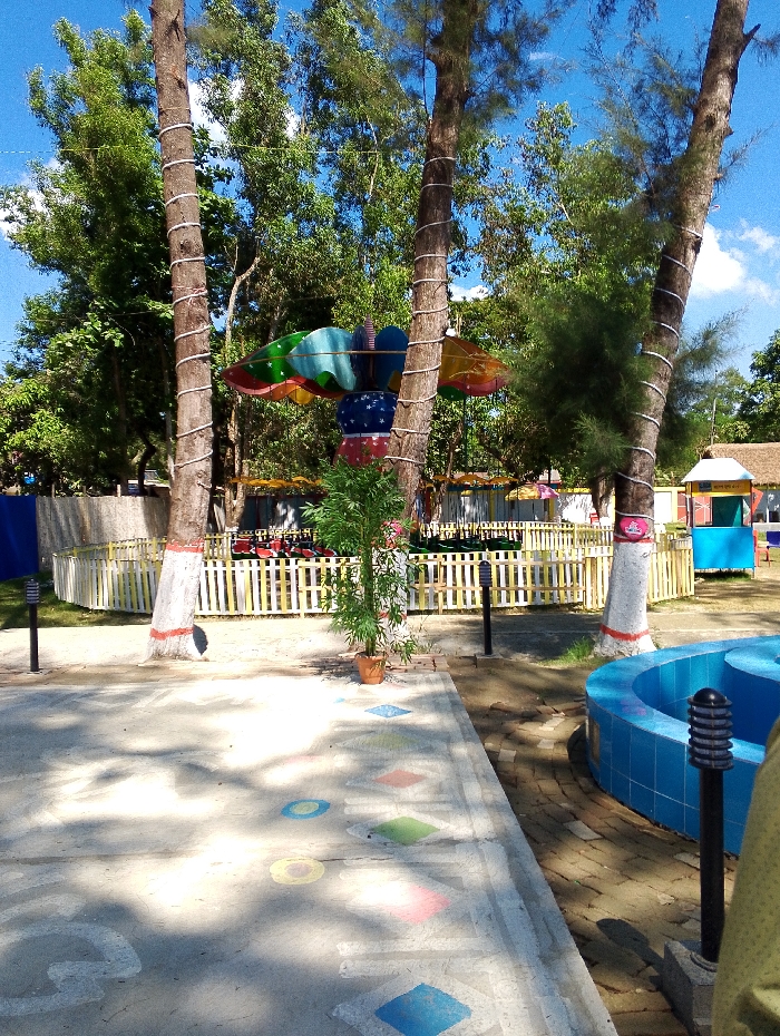 The image shows a playground area in Bhola, Bangladesh, located at coordinates 22.692 latitude and 90.644 longitude. Two tall trees with rough bark stand on either side of a colorful, stationary, kiddie ride shaped like a butterfly. The ride has a large, round, white and blue base, and the wings are made of a multi-colored material, possibly fabric. The playground is fenced off with a white picket fence. A small bush with green leaves grows in front of the ride. A blue pool with a light blue tile border is located on the right side of the image. The ground is paved with light gray concrete. A light blue, green, and orange hopscotch game is painted on the ground between the pool and the ride. A tall, black light post with a rounded top is located in front of the pool. The sky is clear and blue. The weather is sunny and the light is bright. The area appears to be an open-air playground.
