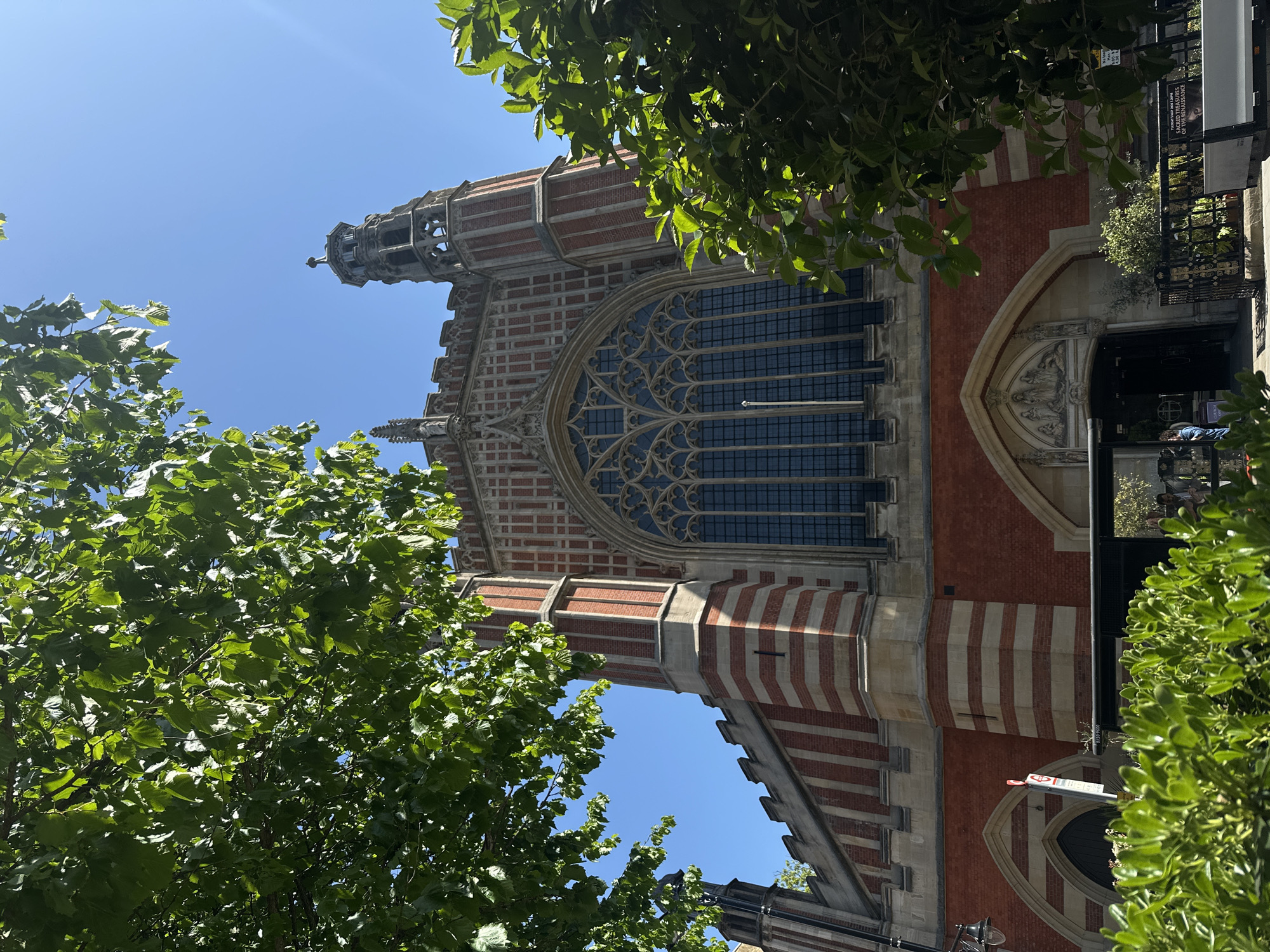The image shows the exterior of the Founder's Building at Royal Holloway, University of London, located in Egham, Surrey, near London, United Kingdom. The building is depicted from a low angle, looking upwards towards its elaborate Gothic Revival architecture. The façade features red brickwork with stone banding and pointed arch windows with intricate tracery. The structure also has a tower with architectural detailing. The foreground consists of foliage from trees, partially obscuring the view of the building. The sky is visible in the upper part of the frame. A sign is also visible on the right.