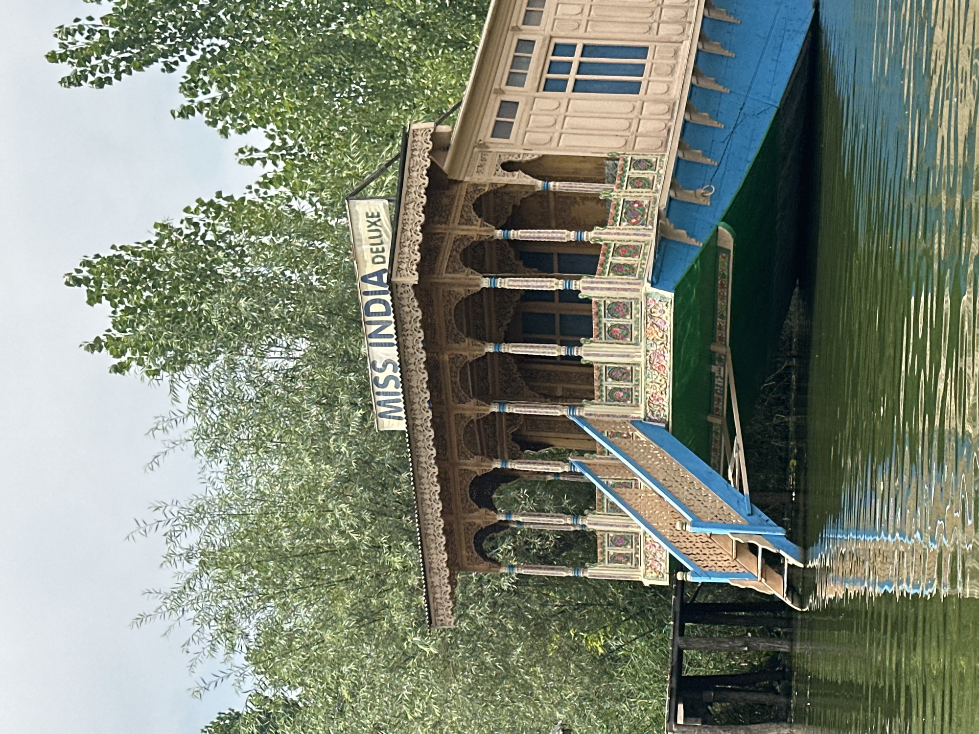 The image shows a brightly colored houseboat named "MISS INDIA DELUXE" docked on a body of water, likely a lake in Srinagar, India. The houseboat is elaborately decorated with intricate woodwork and painted details. A blue staircase with carved railings leads up to the entrance of the houseboat. The water is a murky green, reflecting the trees and the houseboat itself. Lush green trees form a backdrop to the scene. The sky is overcast and a pale grey.