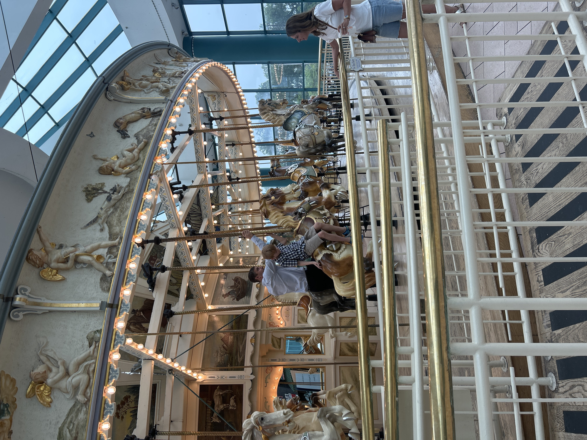 The image shows a carousel in the Great Northern Mall, city of Syracuse, United States of America. A man and a boy are riding on the carousel horses. The boy is wearing a blue and white checkered shirt and khaki shorts, and the man is wearing a light blue dress shirt and black pants. A woman in a white t-shirt and denim shorts is standing by the railing, holding a drink. The carousel is ornate with painted panels and decorative figures. The ceiling of the mall is made of glass, allowing natural light to filter in. The overall scene depicts a family enjoying an outing at the mall.