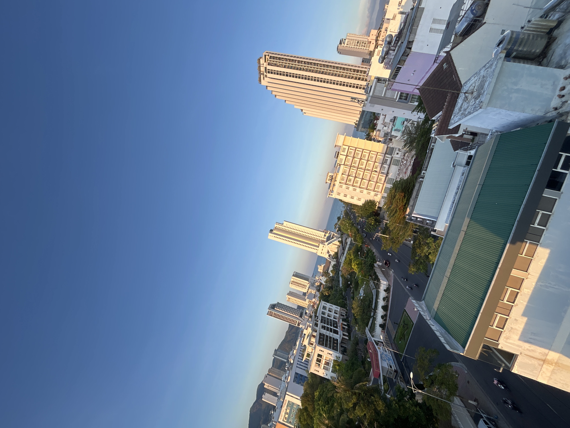The image shows a high-angle view of a cityscape in Bac Nha Trang Ward, Vietnam, featuring a clear blue sky above a variety of buildings, ranging from tall skyscrapers near the coast to smaller structures closer to the vantage point. A road with moderate traffic is visible, lined with trees. The buildings display different architectural styles and heights, suggesting a mix of residential and commercial use. The light indicates it may be early morning or late afternoon.