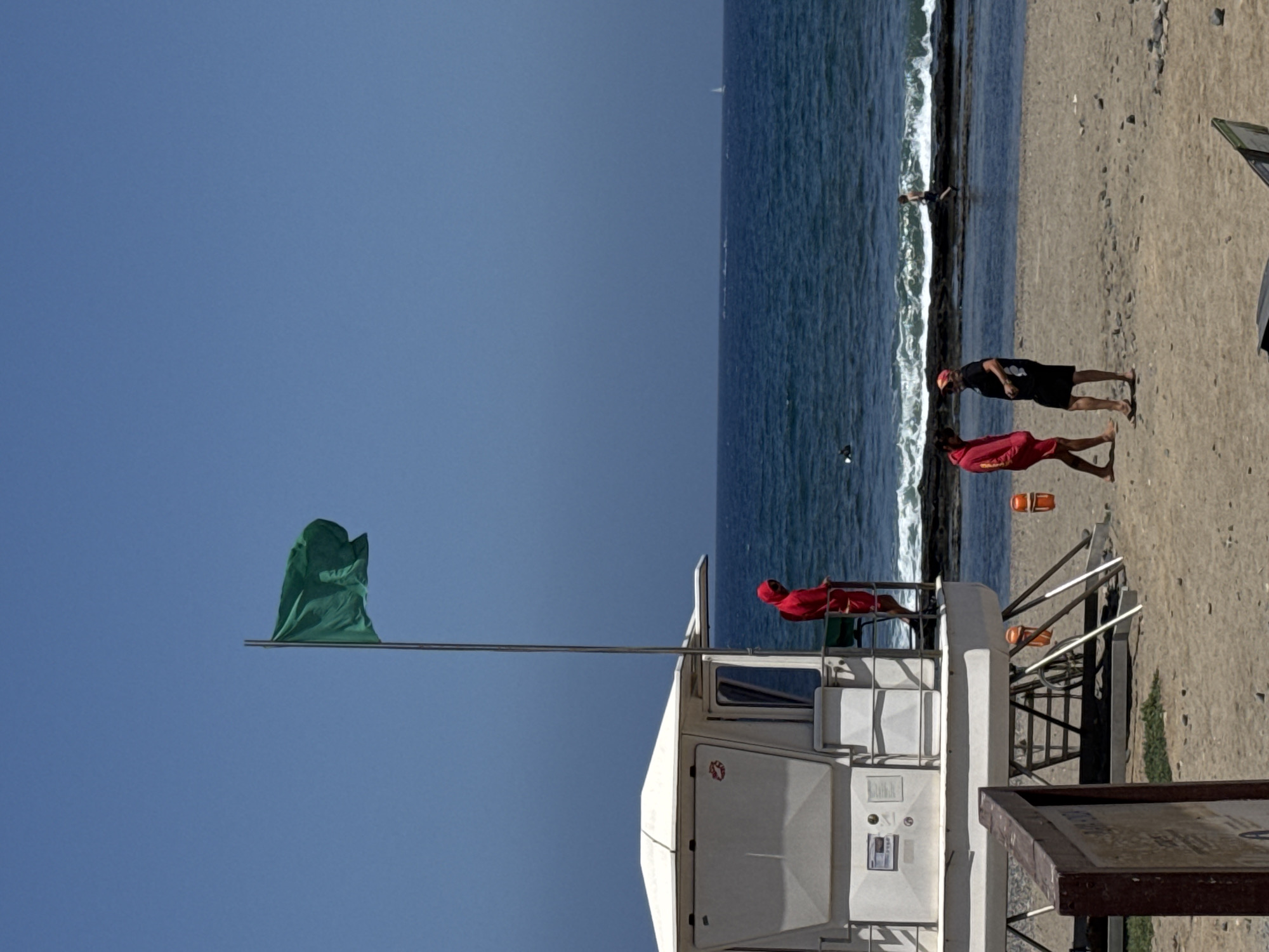 This image depicts a beach scene in Arona, Spain, on a clear, sunny day. In the foreground, a white lifeguard tower stands prominently, with a green flag flying from a pole attached to its side. A person in a red lifeguard uniform is visible on the upper platform of the tower, looking out towards the ocean.
The ocean is a deep blue, with gentle waves breaking on the sandy shore. Further back, two individuals in casual attire are standing on the beach, one in a black t-shirt and shorts, the other in a red top and shorts. Another person is seen lying on the sand near the water's edge.
The beach is sandy and appears to be relatively uncrowded. The time of day is likely midday, given the bright sunlight and clear sky. The overall mood is relaxed and summery. A wooden sign with some text is partially visible in the lower right corner, but the text is not legible. The presence of the lifeguard tower and personnel suggests a public beach where water safety is a priority.