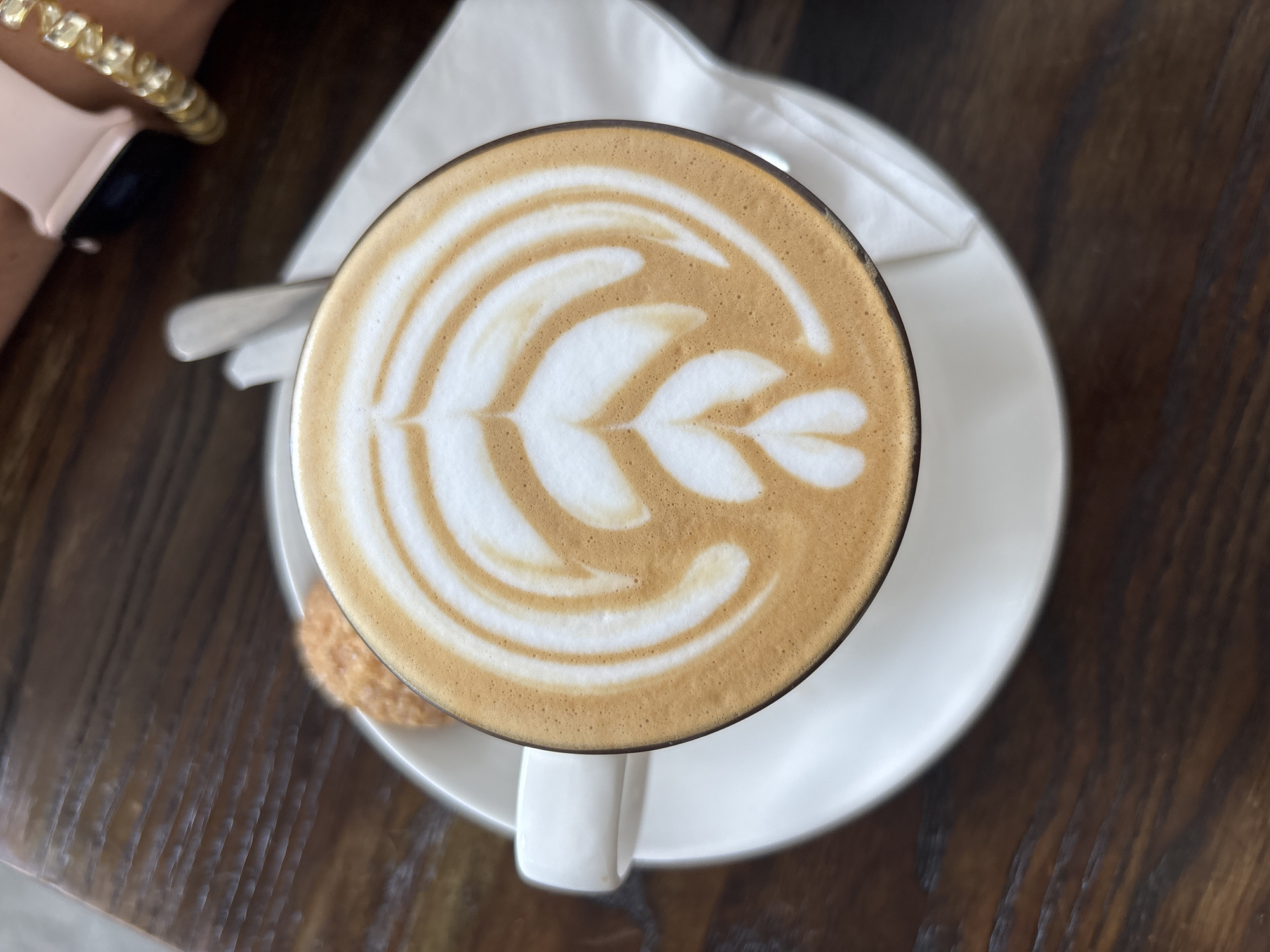 A top-down view of a cup of coffee with latte art on a white saucer is placed on a wooden table. A white napkin and a small biscuit are visible next to the cup. A person's wrist wearing an Apple Watch with a pink band and a gold bracelet is partially visible in the upper left corner of the image. Based on the available information, it is not possible to confirm that this image was taken in the city of Kuta Selatan, Indonesia.