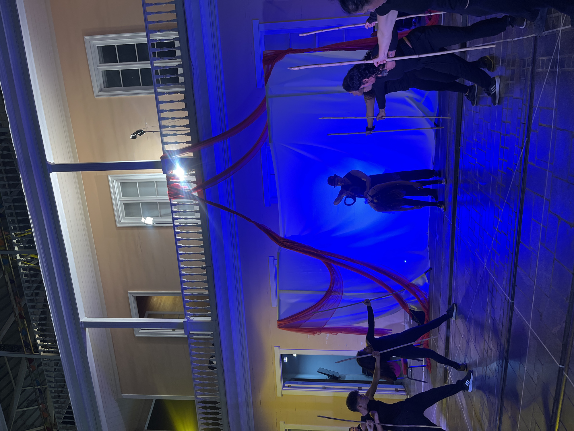This image depicts an outdoor performance taking place at night on a tiled plaza in front of a two-story building with a balcony. The building, painted a peach color, has white trim and columns, and several windows with white frames. The performance area is lit with blue and yellow stage lights, and red fabric is draped as a backdrop.
In the center of the plaza, a performer wearing a dark hat and vest stands next to a donkey, holding a rope. Several other performers, dressed in black, are positioned around the plaza. Some are holding long wooden poles, and appear to be engaged in a coordinated action, possibly part of the performance. One performer on the left is captured mid-motion, leaning forward. Another performer on the right is also holding a pole, with other performers behind them. The overall scene suggests a theatrical or artistic presentation. The architectural style of the building and the balcony suggest it could be a historical structure.