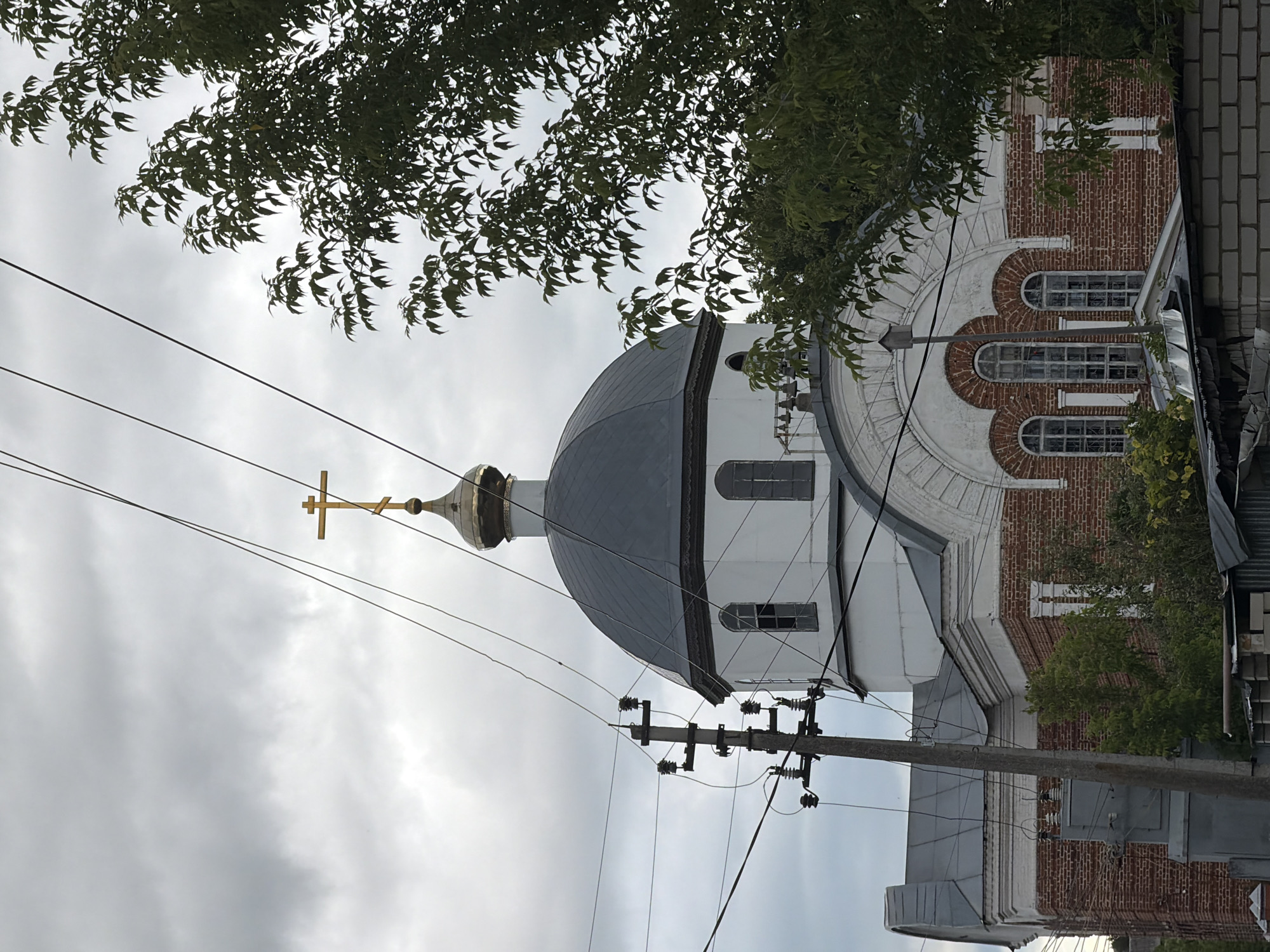 A wide shot captures a cloudy day in an unknown city, focusing on the dome and upper sections of a brick church. The dome is dark gray, topped with a golden Orthodox cross. A weathered wooden utility pole with insulators and wires stands prominently to the left of the church. Thick, dark power lines crisscross the frame, obscuring parts of the sky and the church's architecture. To the right, the dense green foliage of a deciduous tree partially frames the scene. The church itself features red brick lower levels and white stucco upper sections, with arched windows visible. The overall impression is one of a somewhat overcast, ordinary day in a location where urban infrastructure and religious architecture coexist.