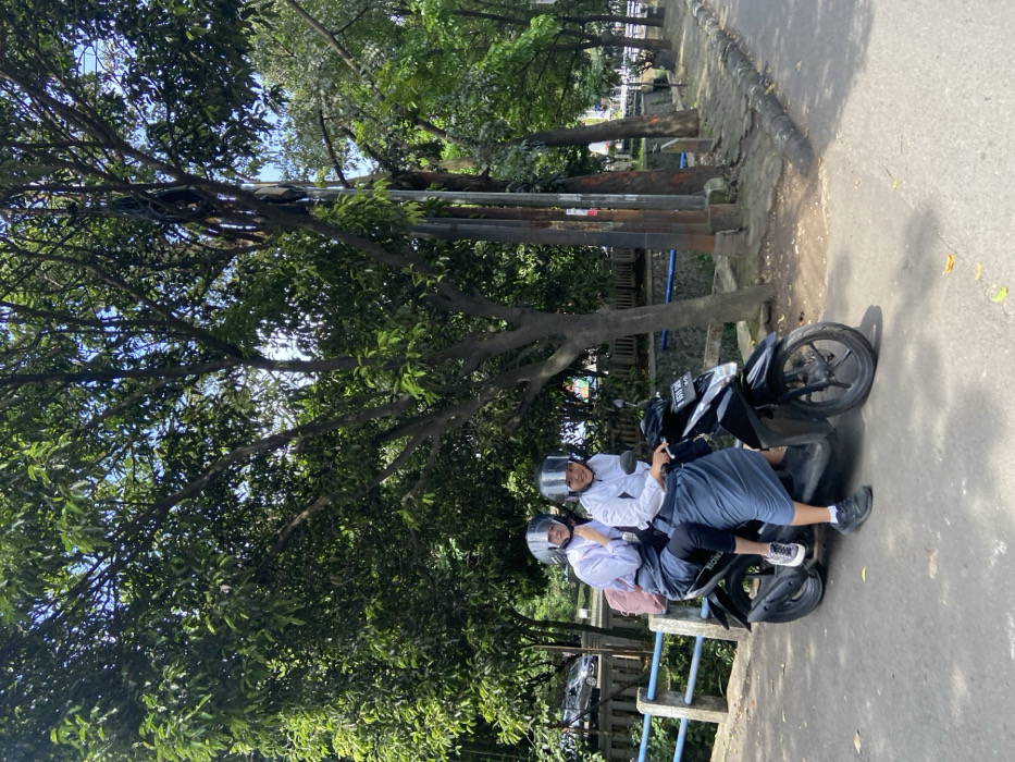 Two young women are sitting on a black scooter. The woman in the front is wearing a white long-sleeved shirt, a white hijab, and a black helmet. The woman in the back is wearing a white long-sleeved shirt, a blue hijab, and a black helmet. They are both looking at the camera. The scooter is parked on the side of a paved road. There is a blue metal railing and a small drainage ditch on the left of the image. On the right side of the image there are trees and bushes. The scene is located in Kecamatan Cimahi Selatan, Indonesia, at the coordinates (latitude: -6.904, longitude: 107.565).