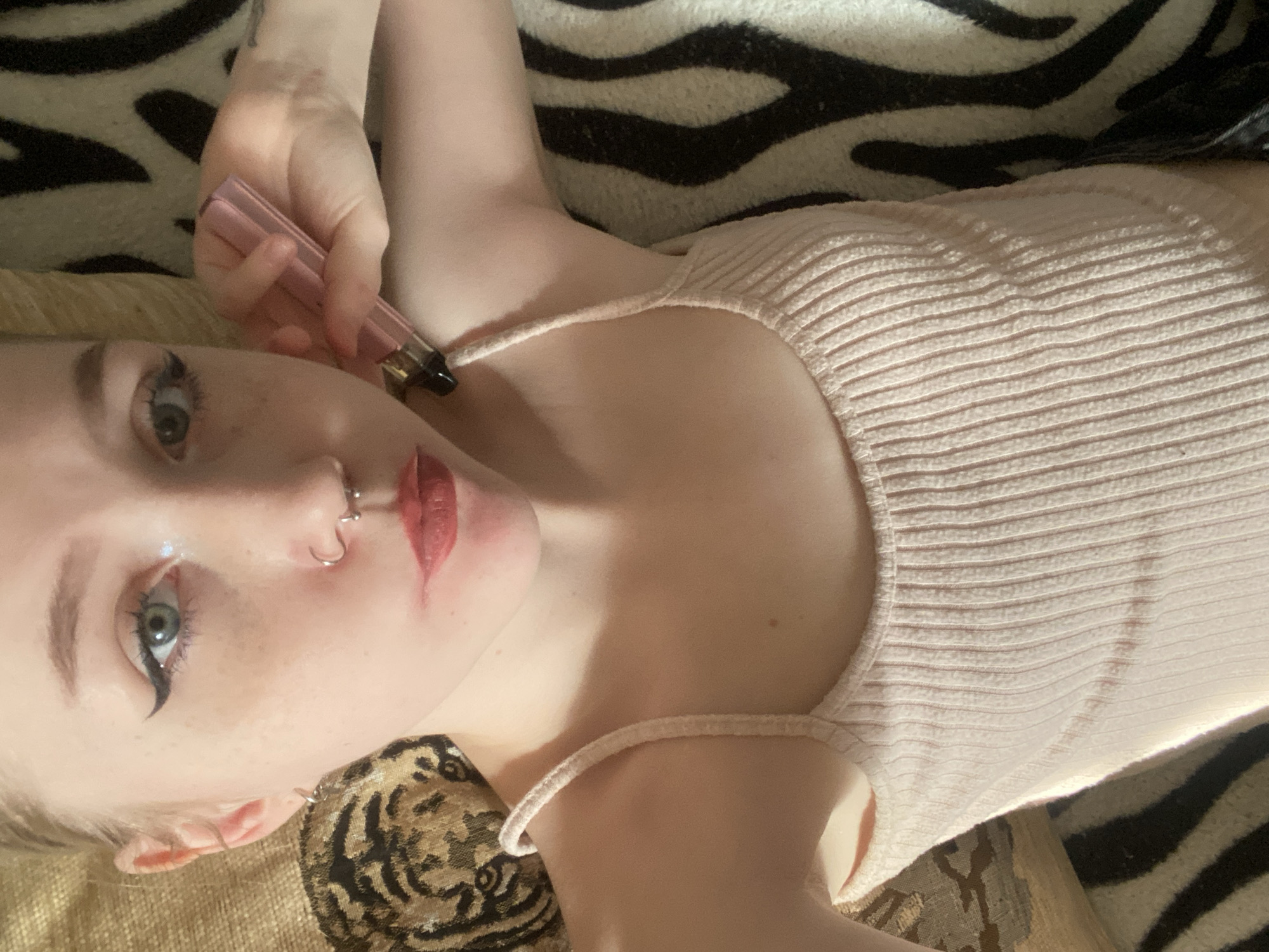A young woman with fair skin, blue eyes, and red lips lies on a zebra-print patterned surface. She is wearing a light pink ribbed tank top. She has a nose piercing and is holding a pink electronic cigarette near her face. The scene includes a patterned cushion beneath her head. It is not possible to determine if the photo was taken in Izhevsk, Russia.