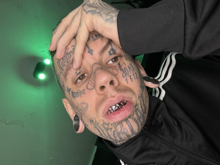 A close-up, head-and-shoulders portrait depicts an individual with extensively tattooed facial features, neck, and visible hands, captured indoors in what is identified as Buenos Aires, Argentina. The subject, with brown eyes, stares directly into the camera. Their head is tilted to the right (viewer's right), and their left hand (viewer's right) rests against their temple, fingers slightly splayed. The visible facial tattoos include various script forms, abstract lines, a cross near the left temple, and a "band-aid" design across the nose. The subject's upper teeth are covered by reflective silver-colored grillz, and both earlobes feature large, circular, silver-toned ear gauges. The individual wears a black track jacket with a prominent zipper and white stripes visible on the right shoulder/sleeve area, along with a black ribbed cuff on the left arm. The background consists of a uniformly green-lit wall or ceiling, showing some surface imperfections or peeling paint. Two distinct circular green light sources are visible on the upper wall/ceiling behind the subject's head.