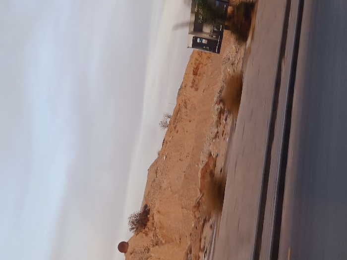 The scene, located in Kafr Sousa Municipality, Syria, captures an outdoor environment under a light grey, overcast sky. The foreground shows a dark asphalt road with a lighter grey concrete sidewalk and a dark grey curb running parallel to it. Faint yellow road markings are visible on the asphalt towards the lower left. In the mid-ground, a prominent, unpaved hillside composed of light brown/tan earth and rocks rises, featuring sparse, dry, shrub-like vegetation on its slopes. On the crest of this hillside, towards the upper left, a large, dark, spherical object, possibly a storage tank or structural element, is partially visible. To the right, a large billboard structure stands near the road, displaying dark panels with illegible text, though some Arabic script is faintly discernable. Green foliage is partially visible in front of the billboard. The image exhibits motion blur, particularly in the foreground and right side, consistent with being captured from a moving perspective. No individuals are present in the frame.