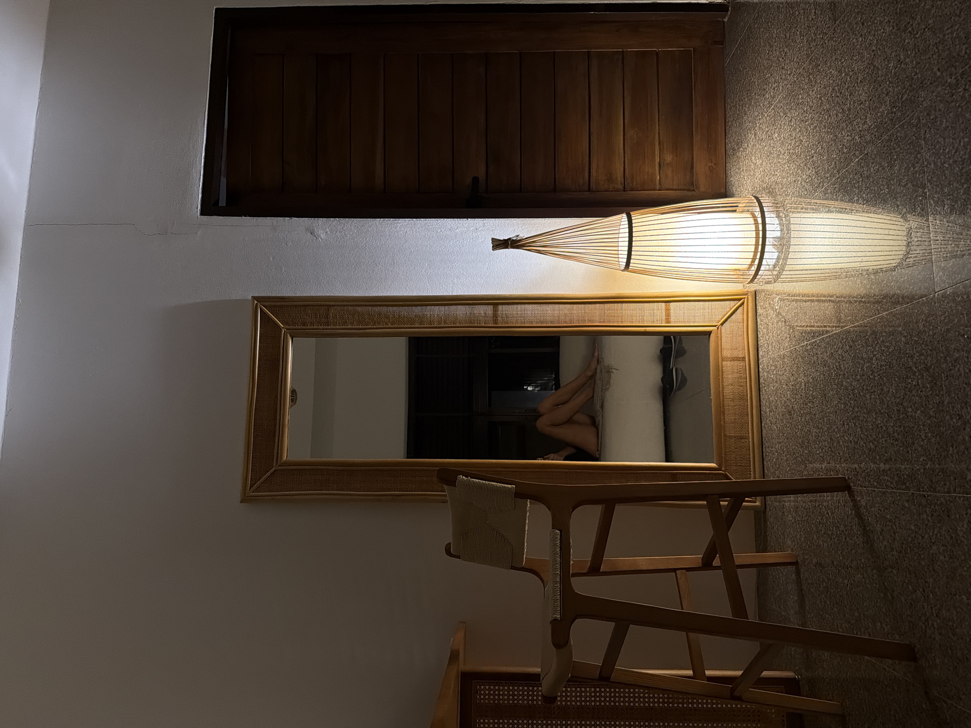 In Baan Bo Phut, Thailand, an indoor scene is captured in low light. A tall, narrow, woven floor lamp with a warm glow illuminates the dark grey, speckled tile floor, reflecting its light. To the left of the lamp is a large, rectangular mirror with a woven wood frame, leaning against a white wall. The mirror reflects a person, only their legs and lower torso visible, sitting on a light-colored surface. Next to the mirror and slightly in front of it is a wooden bar stool with a woven seat and back. The stool is positioned next to a piece of furniture, also with woven accents, that extends out of frame to the left. To the right of the mirror and lamp is a closed wooden door with horizontal planks and a dark handle. The wall above the door shows a thin, vertical crack.