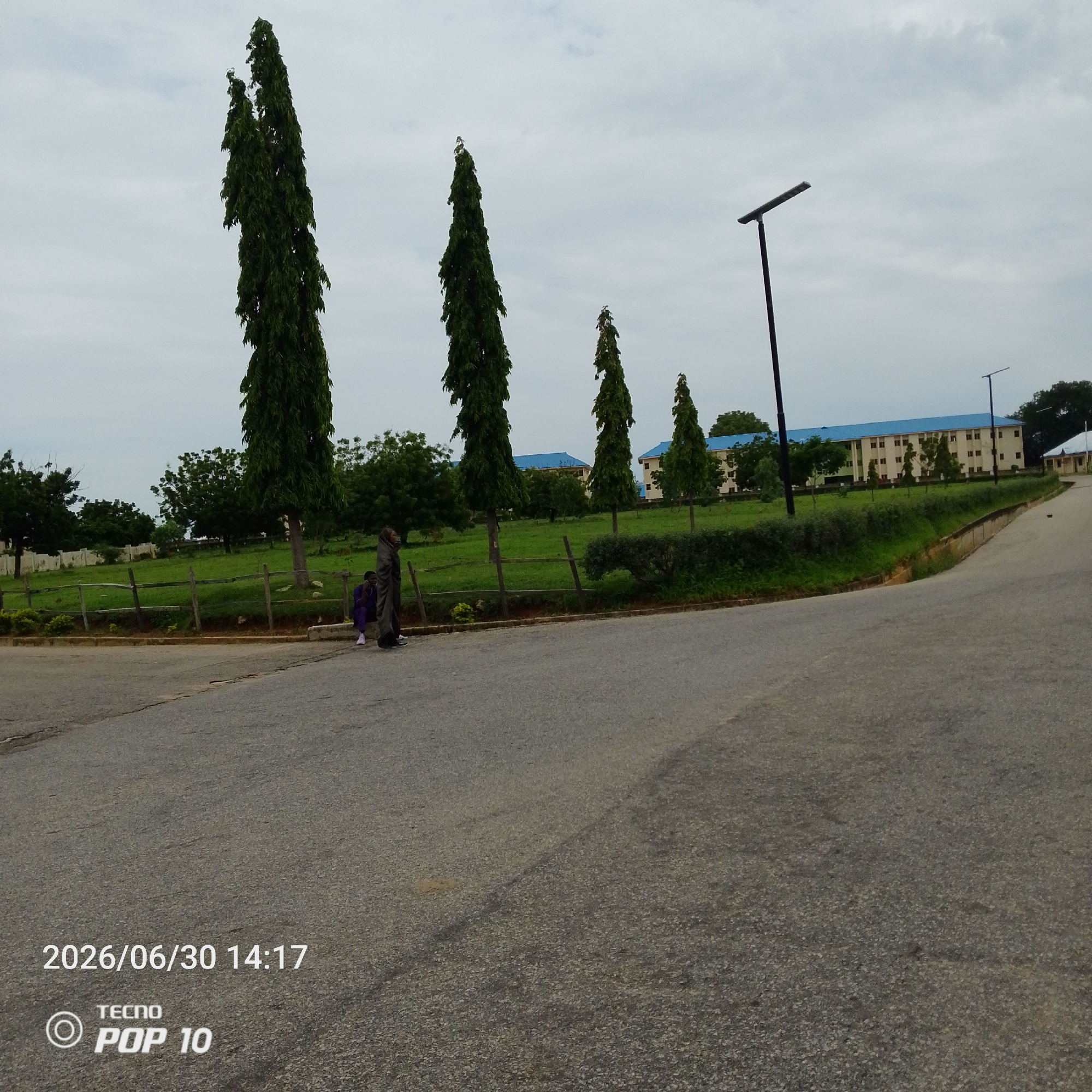 The image depicts an outdoor scene in Jalingo, Nigeria, under an overcast sky, suggesting a cloudy or possibly rainy day. In the foreground, a paved road curves towards the right, with a thin strip of grass and a low wooden fence bordering a larger grassy area. Several tall, slender evergreen trees stand prominently in the mid-ground, their dark green foliage contrasting with the lighter green grass. Behind the trees, a long, multi-story building with a bright blue roof and light-colored walls is visible, hinting at an institutional or educational setting.
Two individuals are standing near the fence in the mid-ground. One person is wearing a dark head covering and a long, flowing garment, and appears to be holding something. Another person, dressed in purple, is seated on a low concrete barrier. Their posture suggests a moment of rest or waiting. Scattered across the grassy area are more trees, some of which are shorter and bushier. A modern-looking street lamp with a flat rectangular light fixture stands tall near the building.
The bottom left of the image contains visible text. It reads "2026/06/30 14:17" indicating the date and time, followed by a logo and text "TECNO POP 10", identifying the device used to capture the photo. The overall mood of the image is calm and ordinary, capturing a slice of everyday life in this Nigerian location.
