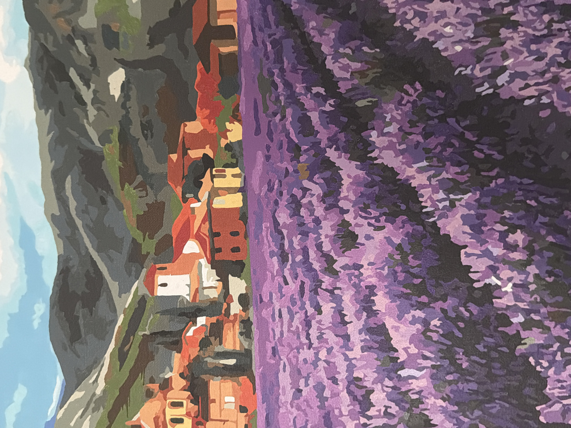 This is a painting depicting a lavender field in front of a village nestled against a mountain range. The sky above is a bright blue with scattered white clouds. The mountains are rendered in shades of gray, green, and brown, with some lighter patches suggesting snow or rock. In the midground, a cluster of buildings with red-tiled roofs and terracotta-colored walls forms a village. Some buildings have white or yellow accents, and one features a white tower with a red cap. The foreground is dominated by a vast field of lavender, painted in various shades of purple, from deep violet to pale lilac, with darker patches of shadow. The style of the painting is somewhat stylized, with visible brushstrokes and a simplified representation of forms, characteristic of a paint-by-numbers artwork. There are no people or animals visible in the scene.