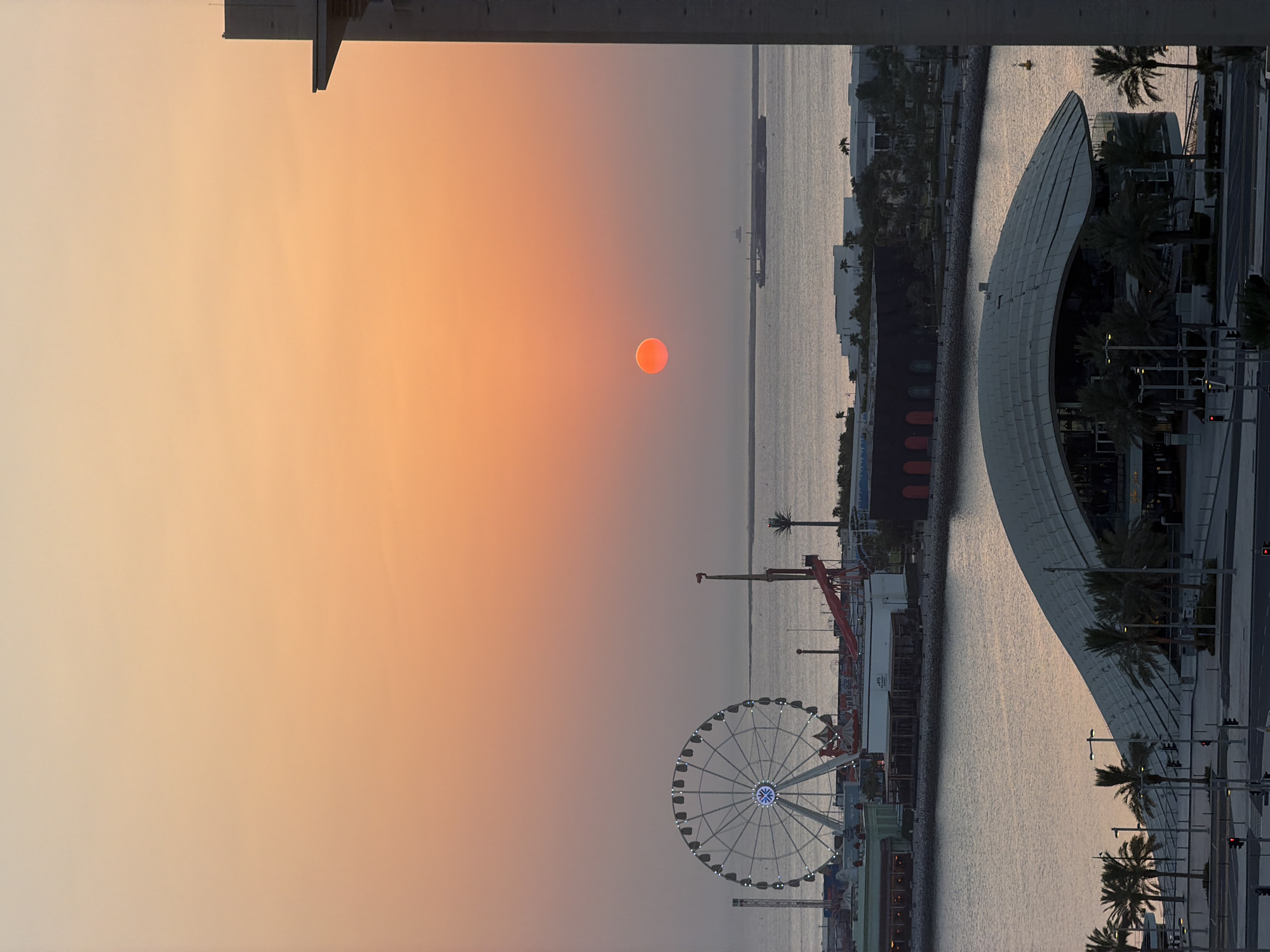 The image shows a sunset over Lusail, Qatar, with a view of the waterfront and the Ferris wheel at Lusail Winter Wonderland. The sky is a gradient of orange and yellow, with the sun a bright red orb low on the horizon. In the foreground, a curved modern building with a white, textured roof is visible, surrounded by palm trees. Beyond this, the waterfront is lined with buildings, including restaurants and entertainment venues. The iconic Lusail Winter Wonderland Ferris wheel stands prominently to the left, illuminated with blue lights. A large cargo ship and a smaller vessel are visible on the calm water in the distance. The right side of the frame is dominated by the edge of a tall, modern building. The scene appears to be a city view at dusk, with an amusement park and waterfront district.