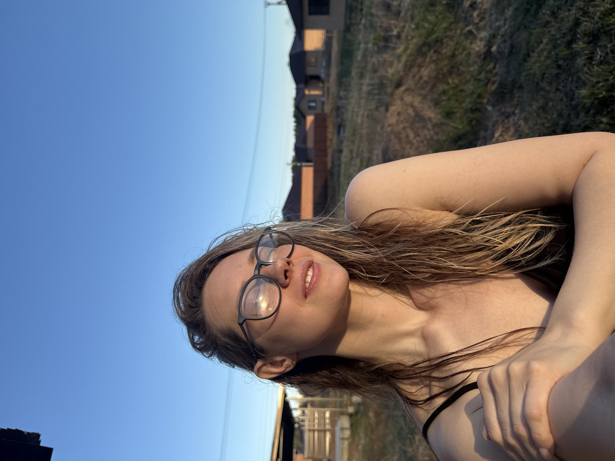A young woman is sitting outside, with a clear blue sky visible in the background. She has long, light brown hair, and wears round-rimmed glasses. The background includes grass and some buildings, possibly residential. The location is likely in Chaikovsky, Russia. The woman's hands are clasped together, and she is looking upwards.