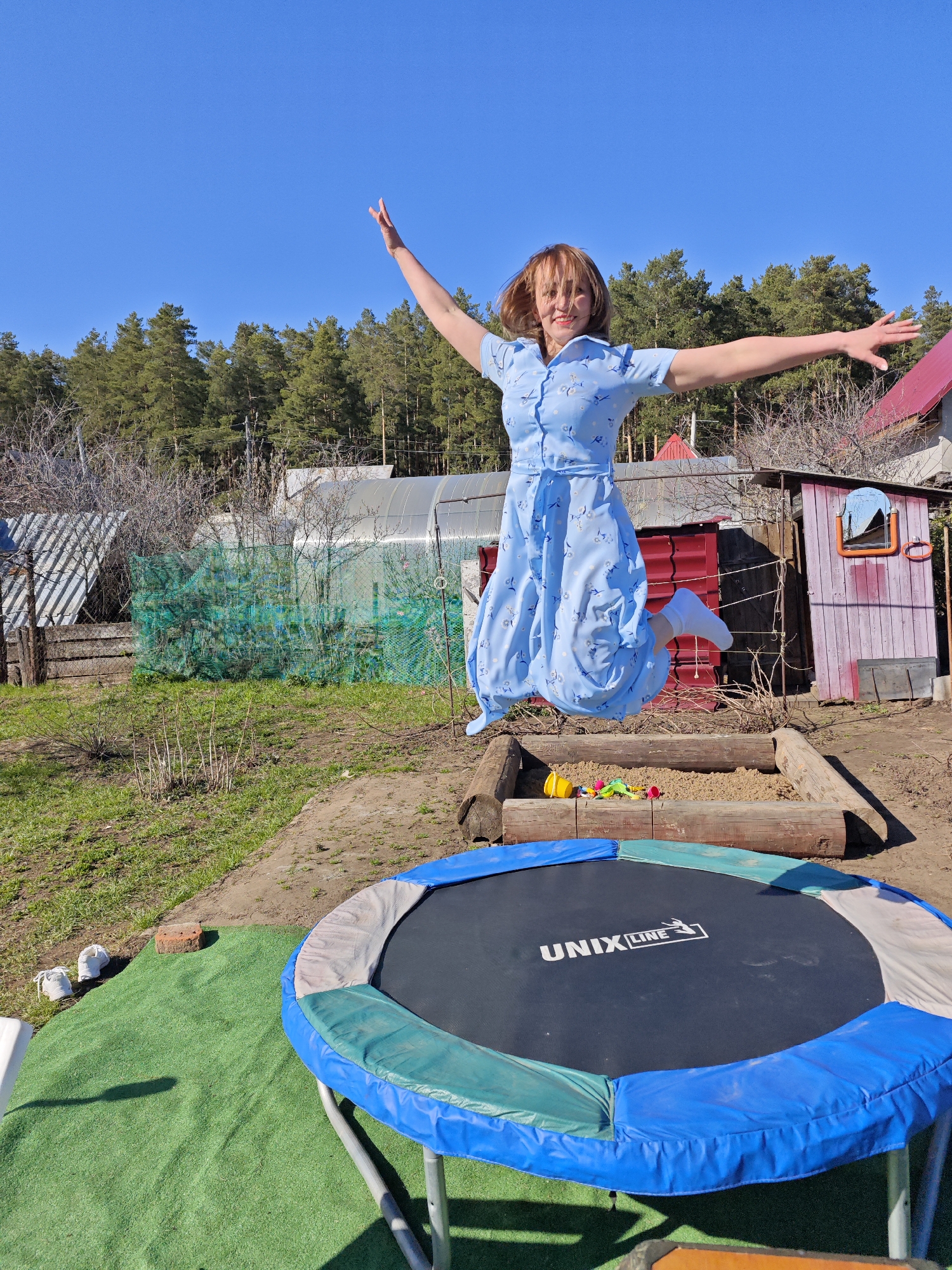 A woman is jumping in the air in a backyard setting. She is wearing a light blue dress with short sleeves and white socks. Her arms are outstretched. Below her is a wooden sandbox filled with sand and colorful plastic toys. A round trampoline with blue padding and the words "UNIX LINE" printed on the black jumping surface is in the foreground. To the left of the trampoline is a patch of artificial green turf with two white shoes resting on it. In the background, there's a shed painted in shades of pink and red, and a greenhouse. The environment consists of a yard with trees and various backyard structures, suggesting a suburban or rural setting in Noviy, Russia. The sky is clear and blue.
