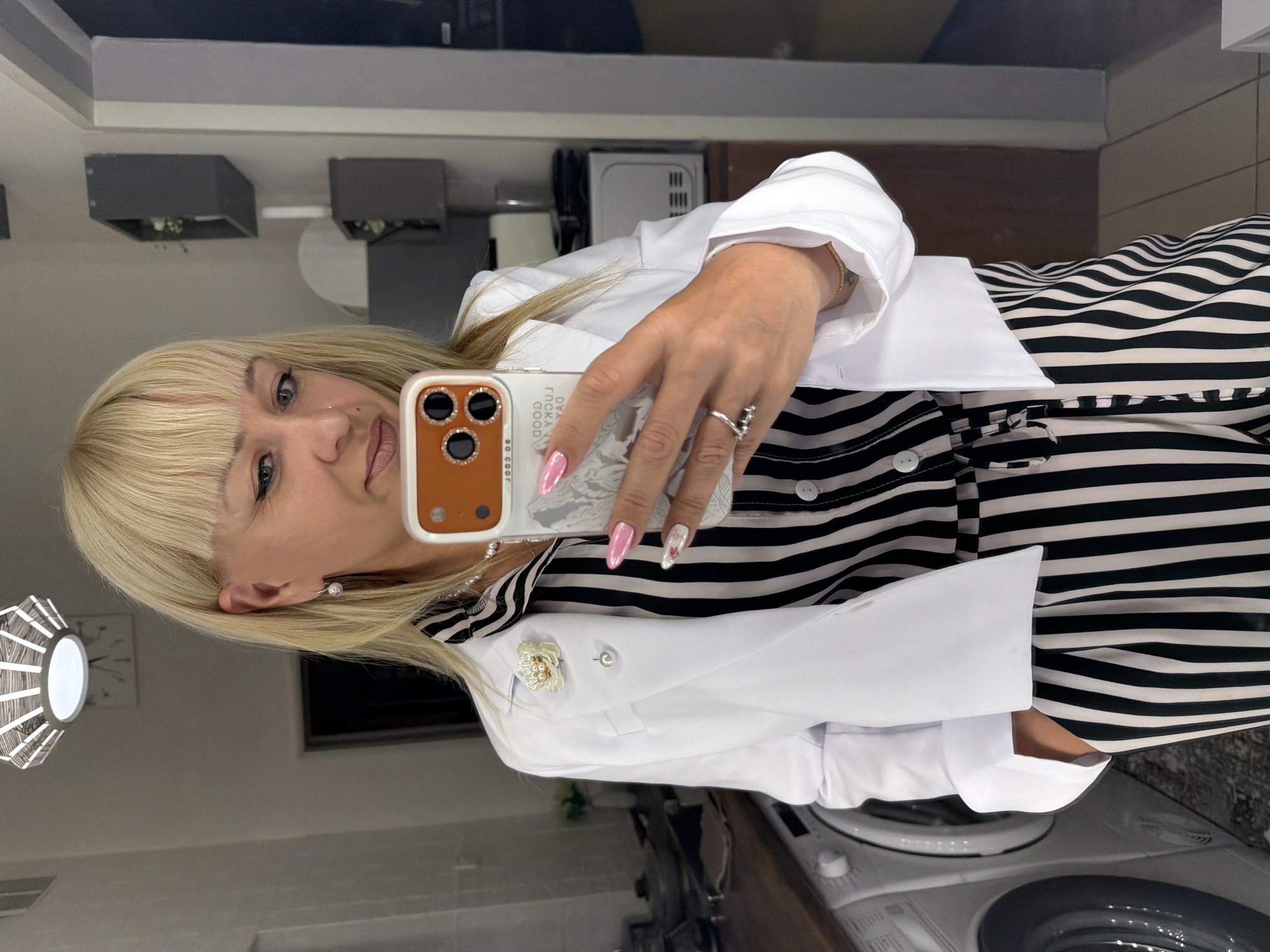 A woman with blonde hair and bangs is taking a selfie in a bathroom or laundry room. She is wearing a black and white striped top and a white blazer with a floral brooch on the lapel. Her nails are painted in a variety of colors and designs. She is holding a smartphone with a brown and white case that says "LUCKY GOOD DAY." The background includes a washing machine, a lamp, and a mirror reflecting the room, including what appears to be kitchen appliances and cabinets. The location might be in the city of Chaikovsky, Russia.
