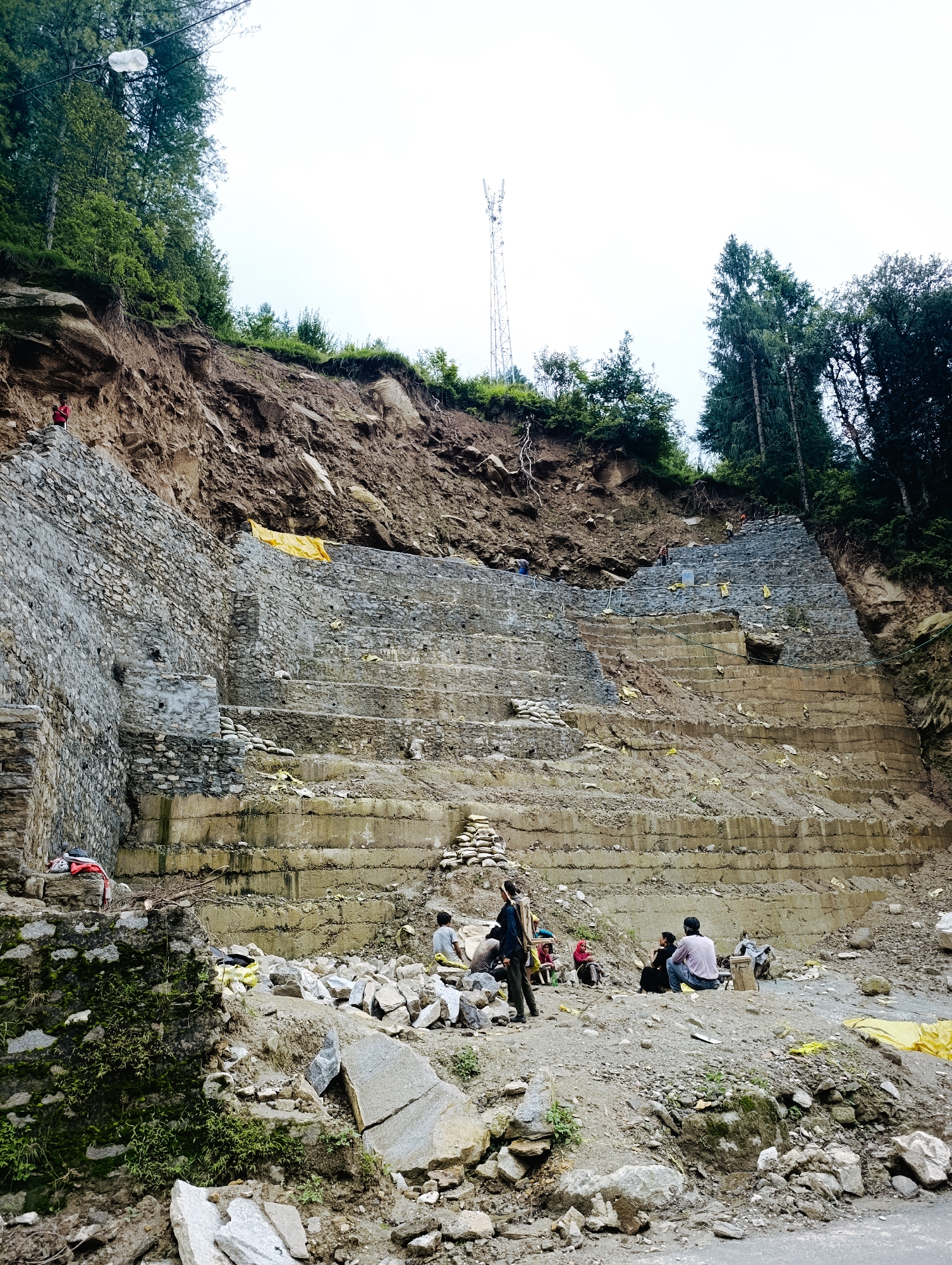 In Vashisht, India, a construction site is depicted on a steep, eroded hillside. The scene is dominated by a massive, stepped retaining wall constructed of rough-hewn stones and concrete, built into the face of a landslide. The wall is tiered with several levels, some reinforced with concrete and others with sandbags. Loose rocks and debris are scattered across the foreground and on the construction levels. Several individuals are present, engaged in various activities. Some are working on the construction, while others appear to be taking a break, sitting or standing among the rubble. A tall telecommunications tower is visible on the ridge above the construction site, along with a dense growth of green trees lining the upper slopes. The sky is overcast, suggesting a cloudy day.