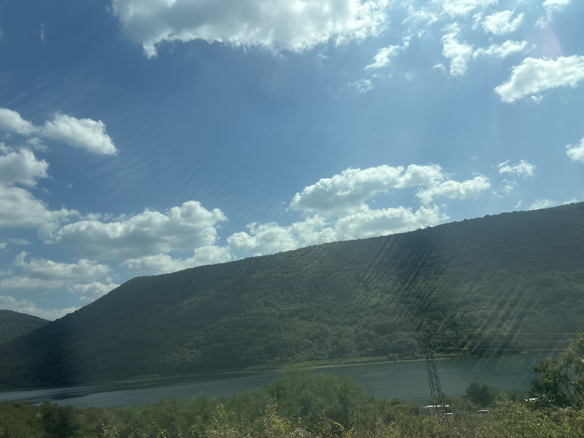 The image depicts a scenic view of a reservoir with a forested hill in the background under a bright blue sky dotted with white clouds. The water is calm, reflecting the sky and the surrounding greenery. The hill is densely covered with trees, creating a lush green landscape. Sunlight filters through the clouds, casting diagonal shadows on the hillside. In the foreground, there is a line of trees and shrubs, and a metal pylon with power lines is visible to the right, near the water's edge. The scene is likely a natural landscape with some man-made infrastructure. While the description mentions the city of Asparuhovo, Bulgaria, this specific visual information cannot be confirmed from the image alone.