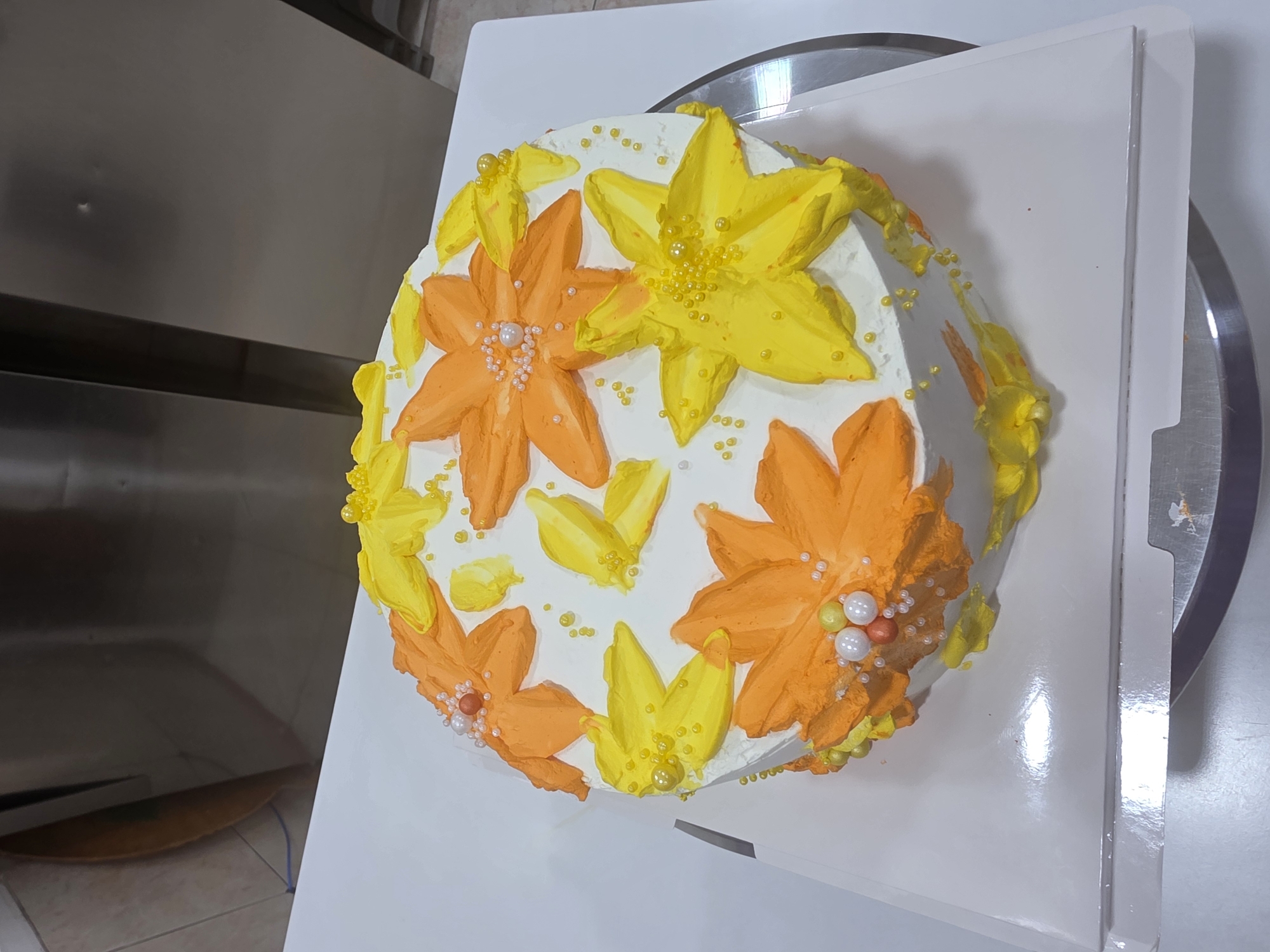 A round cake with white frosting is decorated with orange and yellow buttercream flowers and leaves. Several small yellow and white pearl-like candies are scattered on top and around the flowers. The cake is sitting on a white cake board, which is on a silver turntable. The background shows a metallic surface, possibly part of kitchen equipment. The scene is brightly lit. The location is identified as Dibba Province, Oman.