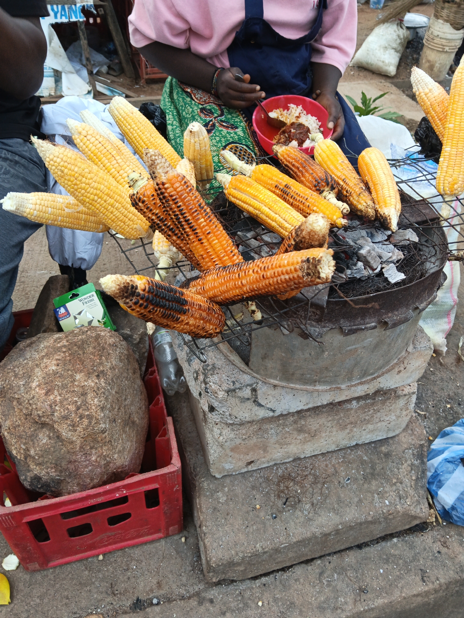 The image depicts a street food vendor in Bukuru, Nigeria, grilling corn. The vendor, dressed in a pink top and blue apron over a green and orange patterned fabric, is seen holding a spoon over a red bowl filled with rice and what appears to be stew. Several ears of corn, some charred from grilling, are arranged on a metal grate over a charcoal fire. In the foreground, a large rock rests in a red plastic crate, and a green packet labeled "Wonder Fresh" is visible. The background shows parts of a market stall, a person in the background wearing a black t-shirt, and various items like sacks and plastic containers. The ground is paved with concrete and dirt, suggesting an outdoor market setting.