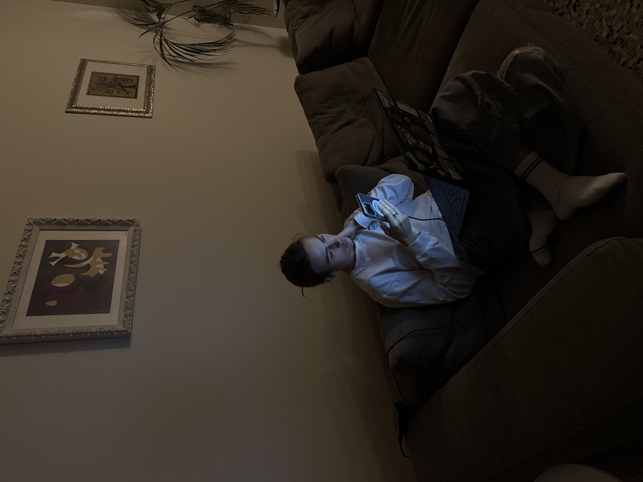 An individual is seated on a dark brown sectional sofa within a dimly lit indoor residence in Venice, Italy. The person, with dark hair pulled back, wears a light-colored long-sleeved shirt, dark trousers with a tear on the left knee, and white socks with blue stripes. They are actively engaged with a brightly illuminated smartphone held in both hands, which is connected by a dark cable. An open laptop, covered with multiple stickers, rests on their lap, its screen also emitting light. The person's left hand is positioned near their neck/shoulder area. The surrounding wall is a neutral light brown. Two framed artworks are visible on the wall: a larger, light-framed piece with abstract purple and gold shapes to the left, and a smaller, gold-framed piece depicting a tree-like design to the right. A tall green potted plant is partially visible behind the sofa on the far right. The floor features light-colored square tiles. The primary illumination in the scene originates from the electronic devices.