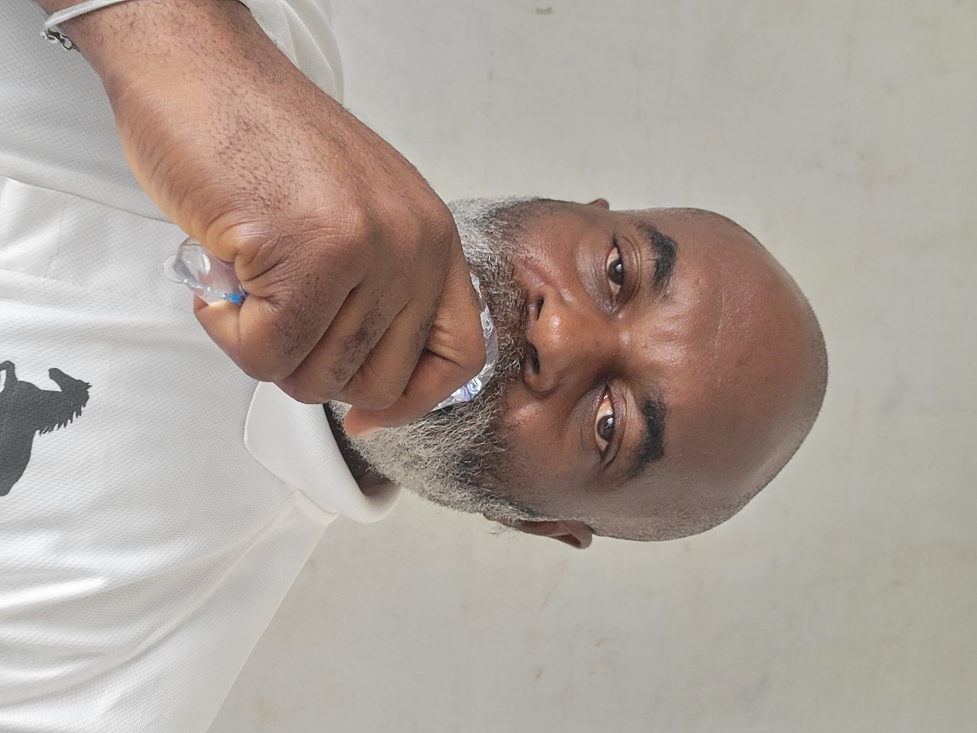 A close-up, tilted shot of a dark-skinned, bald man with a grey beard and mustache, holding what appears to be a toothbrush in his right hand. He is wearing a white shirt with a black design. The background is a plain, off-white wall. There is no information in the image to confirm the location is Maiburiji, Nigeria.