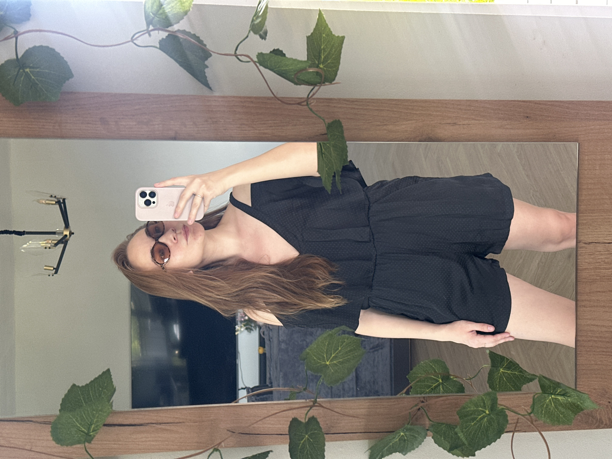 A woman with long auburn hair, wearing brown tinted sunglasses and a black romper, is taking a photo of herself in a mirror. The mirror has a wooden frame with faux vines wrapped around it. The reflection shows a room with a chandelier and a television. The location and city of Kharnavy, Russia are not discernible from the image.