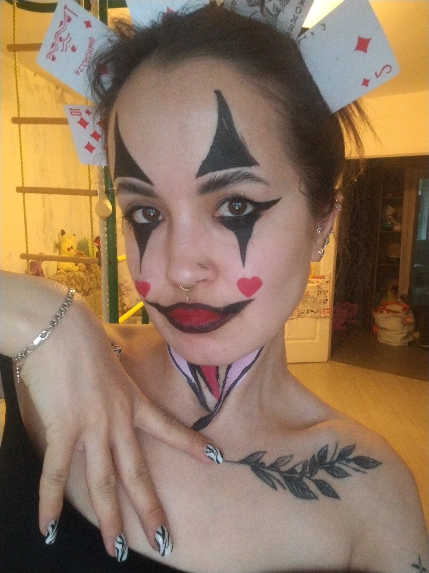 A woman is pictured in a selfie-style image, with clown-like makeup and playing cards affixed to her hair. The makeup includes black diamond shapes around her eyes, red hearts on her cheeks, and dark lipstick. The playing cards are a mix of face cards and number cards. She has a silver bracelet on her left wrist and zebra-striped nail polish. A black leaf tattoo is visible on her right shoulder. A painted diamond-shaped pattern extends from her chin down to her chest. The background shows a room interior, with a wooden climbing structure, toys, and a doorway. The location of the image is not in the city of Noviy, Russia.