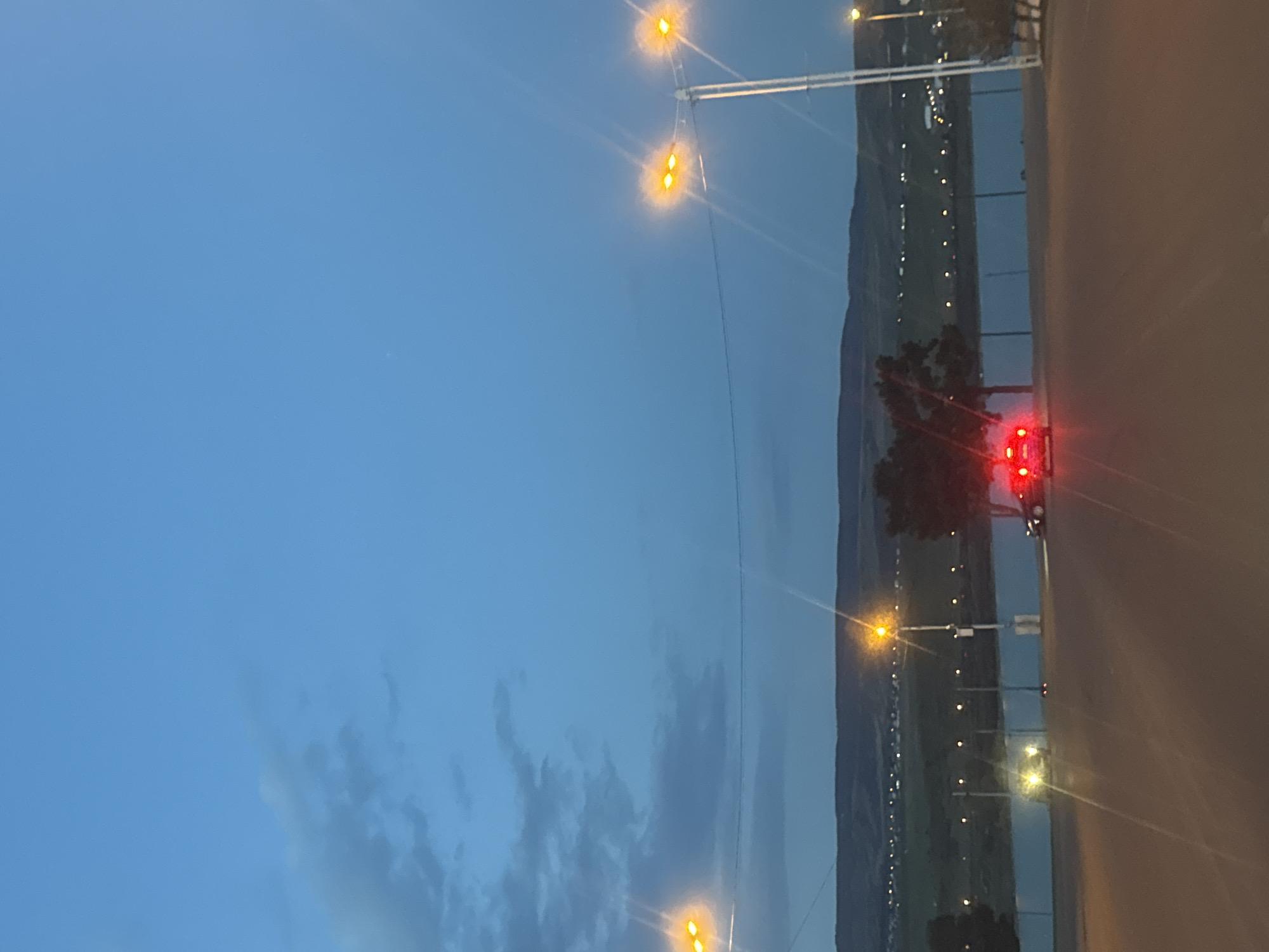 The image, rotated 90 degrees clockwise from its intended orientation, captures a twilight or nighttime scene along a road running parallel to a body of water with mountains in the background.
In the foreground, on the right side of the upright image, a dark road is visible. A vehicle, likely a car, is present on the road, identifiable by its illuminated red taillights, which glow brightly against the dim surroundings. These lights suggest motion or a long exposure, creating a slight streak.
Parallel to the road, a tranquil body of water, possibly a river or lake, stretches across the midground. Beyond the water, a distant landmass or shoreline is dotted with numerous small, bright lights, indicating a settlement or cityscape nestled against the base of dark, imposing mountains that rise against the horizon.
Multiple streetlights with intense yellow-orange glows are prominent throughout the scene. These lights are mounted on tall poles, lining both the road and the edge of the water, casting significant lens flares due to the low-light conditions. Wires are strung between some of the poles.
The sky occupies the upper portion of the image, displaying a deep, rich blue characteristic of dusk or early night. A few faint, wispy clouds are subtly visible against this backdrop. The overall atmosphere is serene and illuminated primarily by artificial light sources. The combination of mountains, water, and artificial lights aligns with the given location context of Tbilisi, Georgia. There is no visible text in the image.