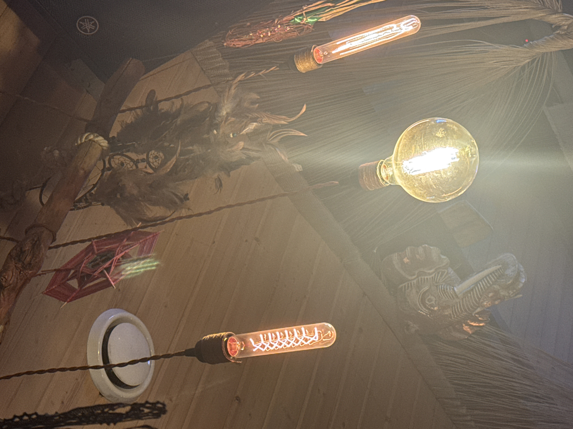 The image depicts an indoor scene with a rustic aesthetic, likely a bar or restaurant interior, in Krasnaya Polyana, Russia. The ceiling is constructed of wooden planks. Several decorative elements are visible, including a circular white light fixture, a red woven geometric ornament, and a collection of feathers arranged as a dreamcatcher. Hanging lights are a prominent feature: one is a tubular bulb with a visible filament, another is a round bulb emitting a warm glow, and a third is another tubular bulb with a similar filament. A carved wooden artifact, possibly an animal head, is mounted on the wall or ceiling. The lighting is warm and somewhat diffused, creating a cozy atmosphere. No people are present in the image. The overall impression is one of a thoughtfully decorated, dimly lit space with a focus on natural and handcrafted elements.