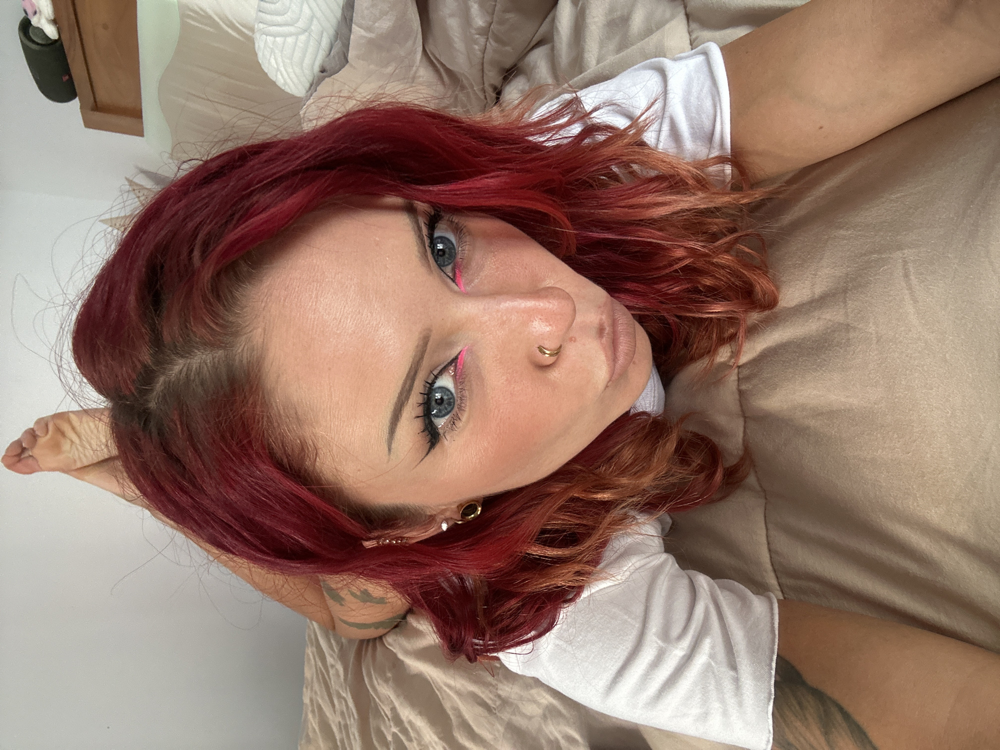 A female with red, shoulder-length hair is lying on a beige bed. She has blue eyes and pink eyeliner. She is wearing a white t-shirt and has a gold nose ring and gold earrings. A portion of a tattoo is visible on her right arm. The background includes a wall and a portion of a wooden headboard. The location is not identifiable from the image.