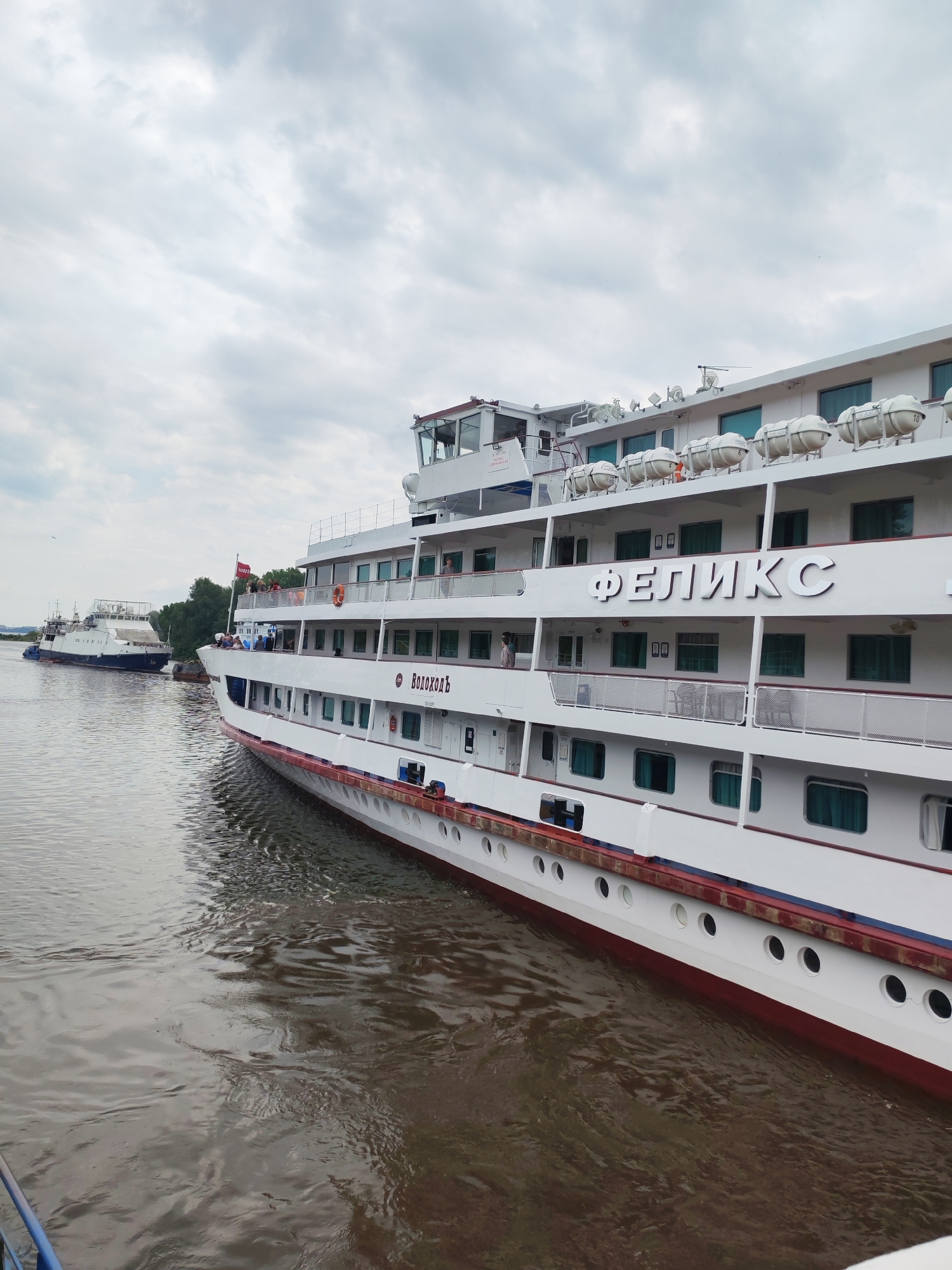 A large white cruise ship named "ФЕЛИКС" is docked on a wide, murky river under an overcast sky. The ship has multiple decks, each with rows of green-tinted windows. Life preservers are visible on the upper deck. The name "ФЕЛИКС" is emblazoned in large, white, Cyrillic letters on the side of the ship. Further down the river, a smaller ferry is visible. The riverbank in the distance is lined with trees, and the sky is filled with soft, grey clouds. The water shows ripples and reflections from the ship and the sky.