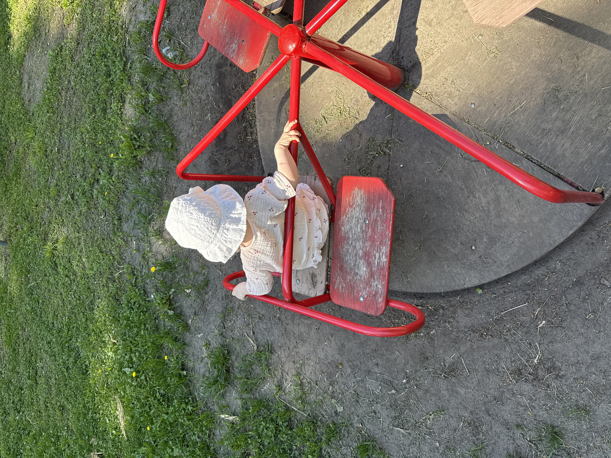 A child wearing a white patterned shirt and a white hat is seated on a red playground merry-go-round in a park in Красный Маяк, Russia. The merry-go-round has a circular gray platform around its base, partially surrounded by grass. The child is holding onto the center pole of the merry-go-round. The photo is taken from above.