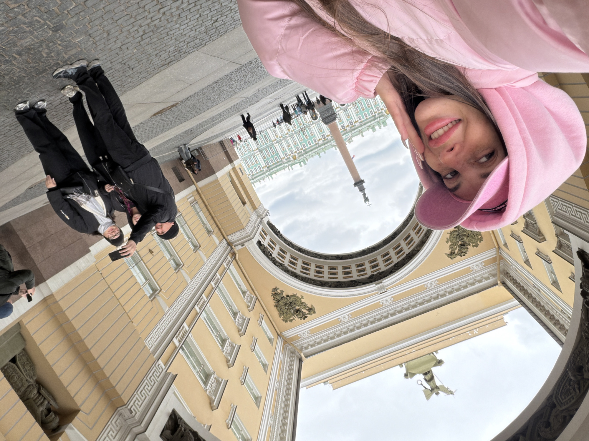 The image, taken with a wide-angle lens and presented sideways, captures a dynamic low-angle view of Palace Square in Saint Petersburg, Russia. The perspective is from under a grand, curving archway of a yellow neoclassical building, looking out into the expansive square.
The primary subject is a young woman in the foreground, taking a selfie. She wears a light pink hooded jacket with the hood up and a matching baseball cap. Her long brown hair frames her face, which is adorned with light freckles. She smiles warmly, looking directly at the camera (her phone), with her right hand gently touching her cheek, revealing long, light-colored fingernails.
In the mid-ground, to the left, two individuals are visible on the paved ground, dressed in black jackets and trousers with sneakers. One of them holds a smartphone, seemingly photographing the surroundings. Further in the distance, more indistinct figures indicate other visitors or tourists.
The architectural setting is magnificent. The yellow building in the immediate foreground features intricate white cornices, window frames, and decorative patterns. The underside of the archway displays ornate white and gold/bronze relief embellishments, including a prominent central emblem. Beyond this archway, the iconic pale green and white facade of the Winter Palace (Hermitage Museum) is clearly visible, stretching across the background. In front of the palace, the tall, slender Alexander Column stands prominently. A statue, possibly an equestrian figure, is also partially visible on a pedestal against the overcast sky in the lower right portion of the scene.
The sky is overcast with a uniform grey cloud cover, suggesting daytime without direct sunlight. The overall atmosphere appears calm, and the main subject's smile conveys a sense of enjoyment during her visit to this historic landmark. There is no visible text in the image.