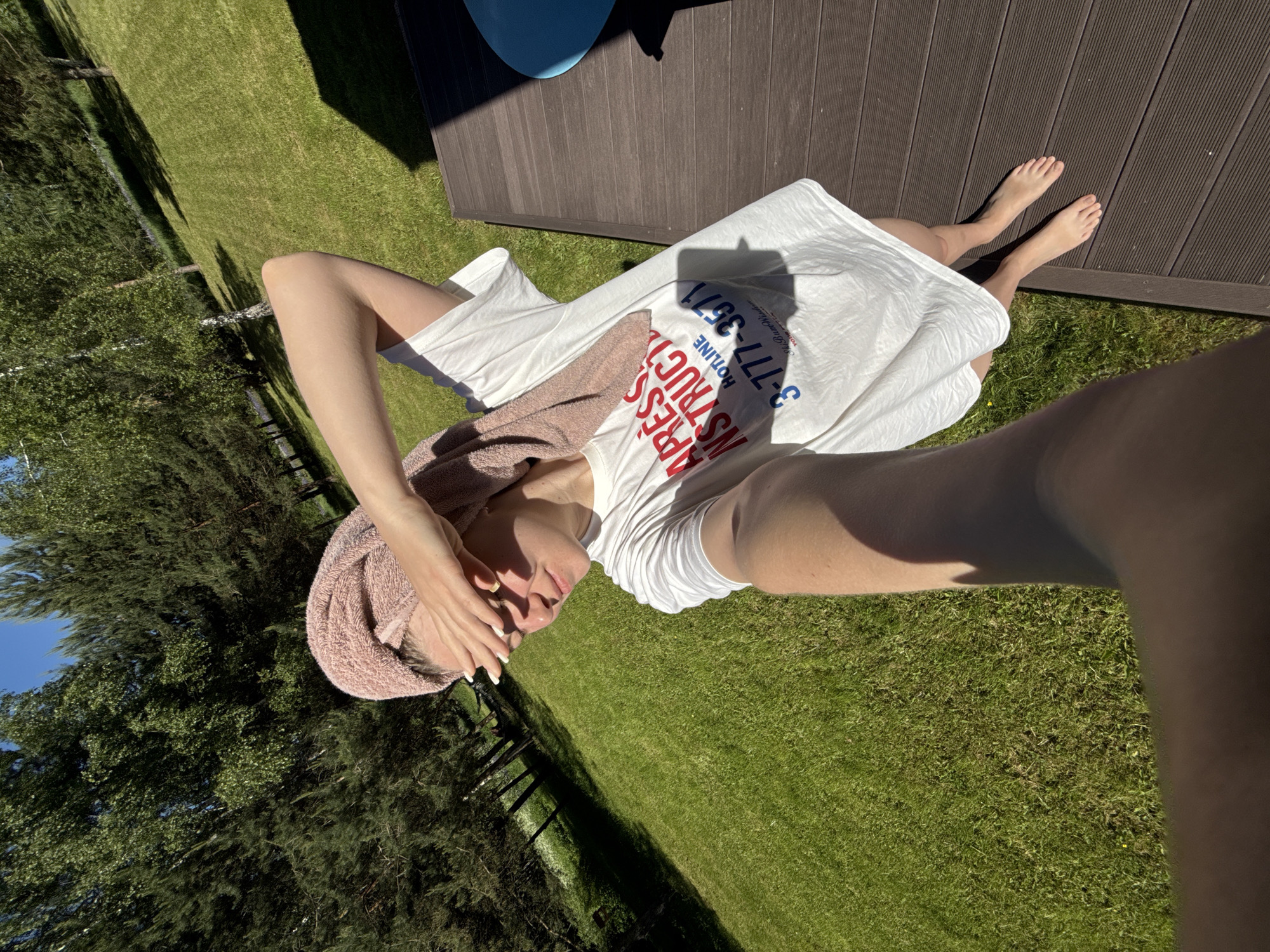 A young woman is depicted in a full-body selfie, standing barefoot on a dark brown slatted wooden deck under bright, direct sunlight. She is wearing an oversized white t-shirt with red and blue text, partially visible, that includes "APRESS INSTRUCTIO," "HOTLINE," "3-777-3571," and "Ski Bum Wisdom." A pink terry cloth towel is wrapped around her head. Her left hand is raised to shield her eyes from the sun, casting shadows on her face. Her right arm is extended, holding the camera. The background consists of a lush green grassy lawn that gently slopes upward, populated by numerous tall trees, including distinct birches and dense conifers, under a clear blue sky. Strong shadows are cast across the deck and grass, indicating intense sunlight. A small, partially visible blue object is situated on the far right of the deck. This scene is located in Копнино, Russia.