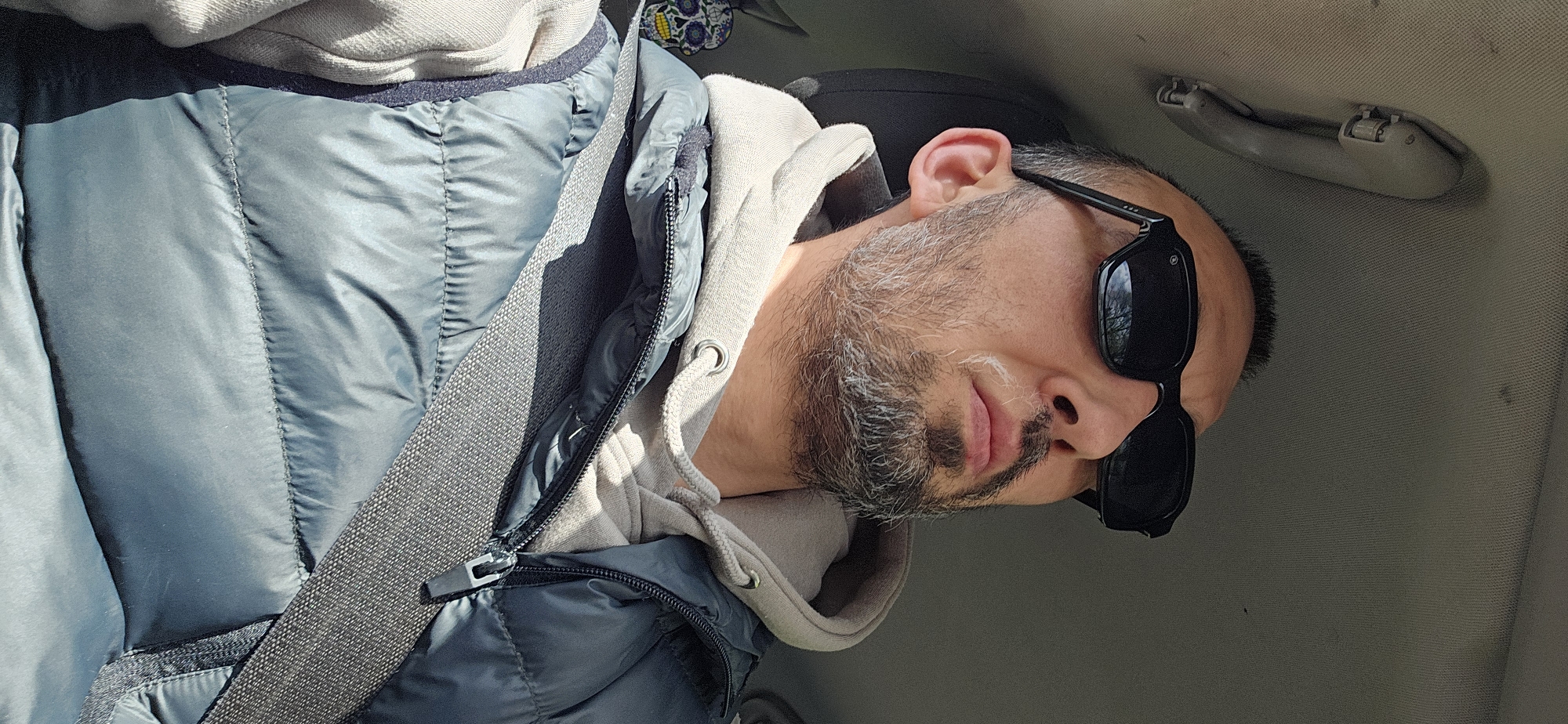An upright-oriented image features a man in a car, likely a passenger. He is seen from the chest up, wearing a light grey or beige hooded sweatshirt layered under a teal or blue-green puffer vest. A grey seatbelt diagonally crosses his chest from his left shoulder.
The man has a beard and mustache, showing noticeable streaks of grey hair. He wears black, rectangular sunglasses that obscure his eyes. His expression appears neutral and relaxed. Bright sunlight illuminates the scene, suggesting it is daytime and possibly sunny outside.
The car's interior is visible, with a light grey or beige fabric headliner on the ceiling. An overhead grab handle, matching the interior color, is mounted above and to the right of his head. No visible text is present on his clothing, accessories, or the car's interior. The setting is a casual car journey, taking place in Norwich, United Kingdom.
