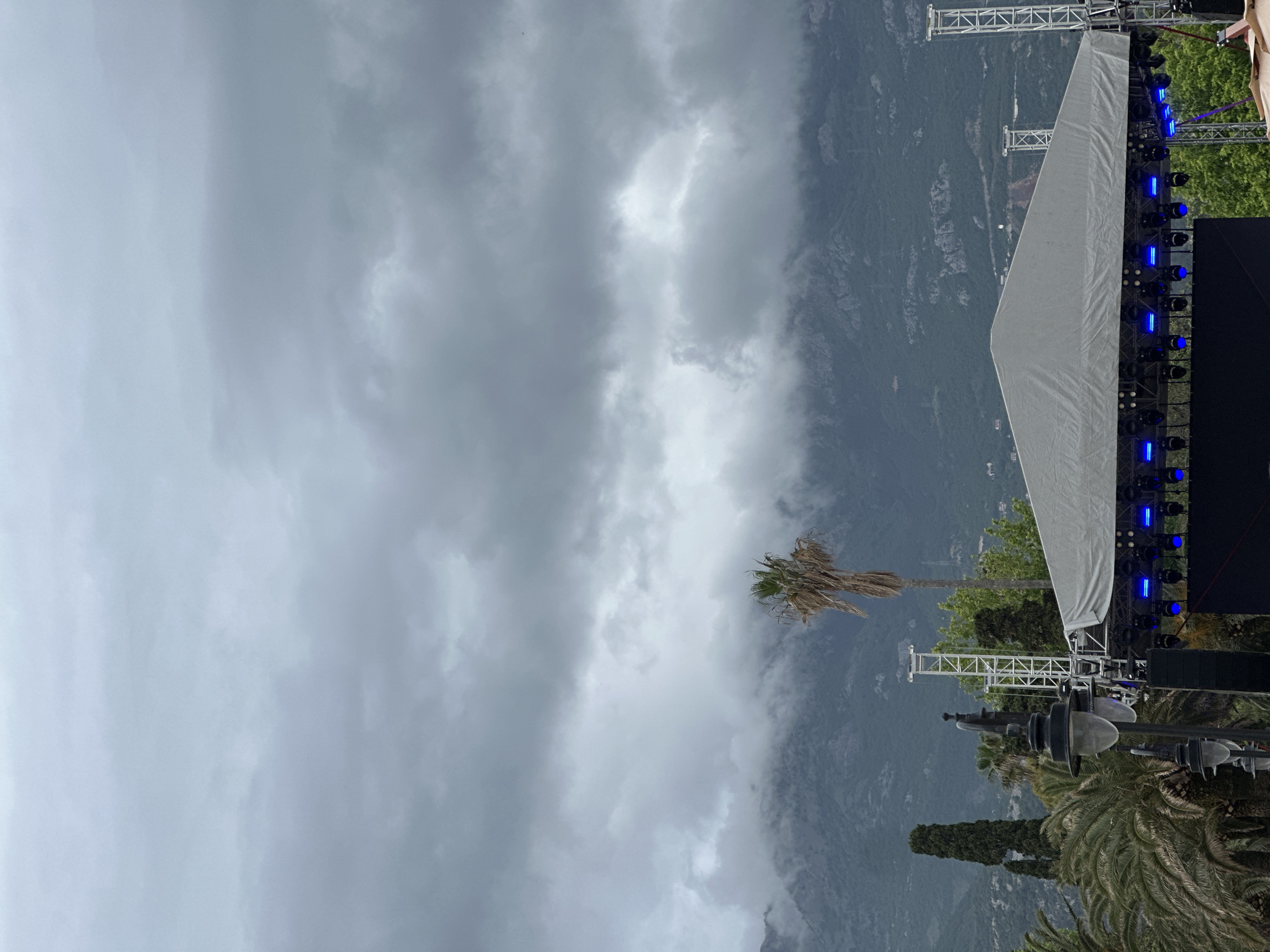 The image shows a cloudy day in Budva, Montenegro. A large outdoor stage is set up, likely for an event. The stage has lighting and a white backdrop or screen. In the background, mountains are partially obscured by clouds. Palm trees are visible near the stage. The perspective is somewhat tilted. There are no visible people in the image.