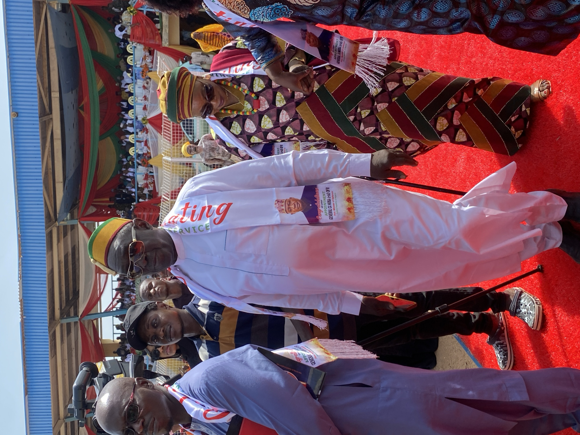 The image shows a group of people in Takau, Nigeria, likely at an event. The foreground features several individuals dressed in traditional attire, wearing sashes that seem to commemorate the appointment of "GENERAL CG MUSA (rtd) OFR". One man wears a white robe and a matching sash with the words "SERVICE" and "ating" visible. Another wears a patterned robe and hat. A red carpet covers the ground. In the background, spectators are visible, seated in rows under a structure decorated with flags or banners in red, green, and yellow. The sky is light blue.