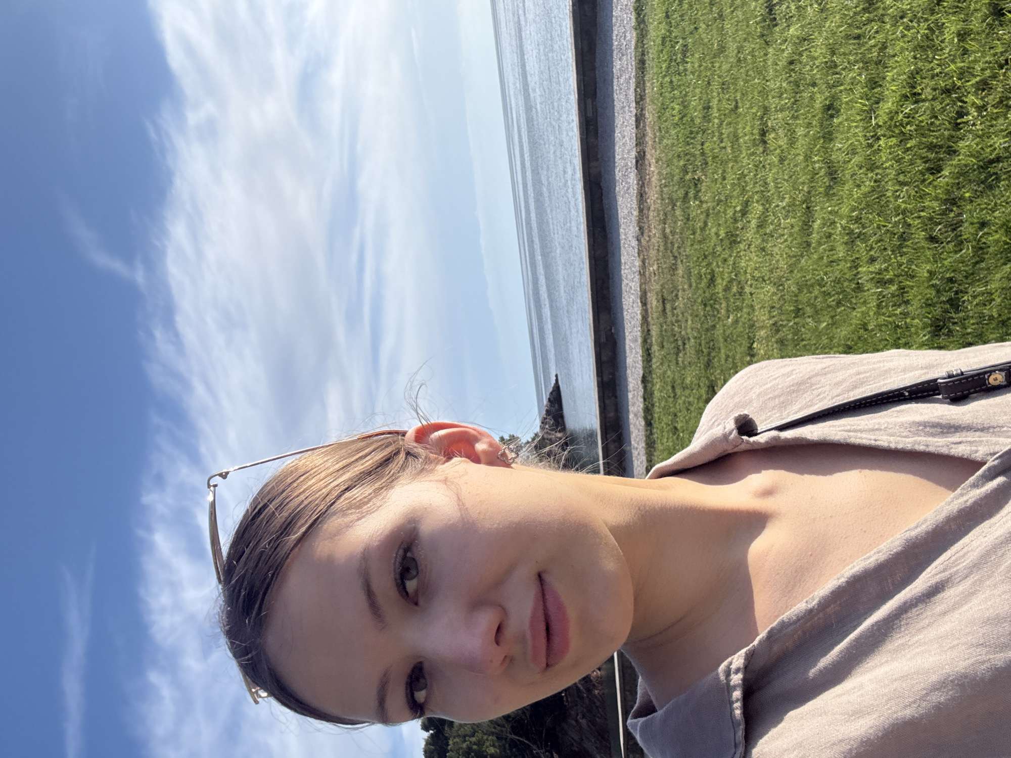 A medium-shot photograph, rotated 90 degrees counter-clockwise, shows a woman's face in the foreground, with a landscape in the background. The woman has fair skin, brown hair pulled back, and a small earring in her left ear. Her gaze is directed towards the camera. Sunglasses are partially visible in her hair. She is wearing a light-colored shirt. The background features a blue sky with scattered white clouds. A body of water, potentially the Adriatic Sea, is partially visible, bordered by a stone wall and a grassy area. The scene appears to be taken near Sveti Stefan, Montenegro, based on the landscape.