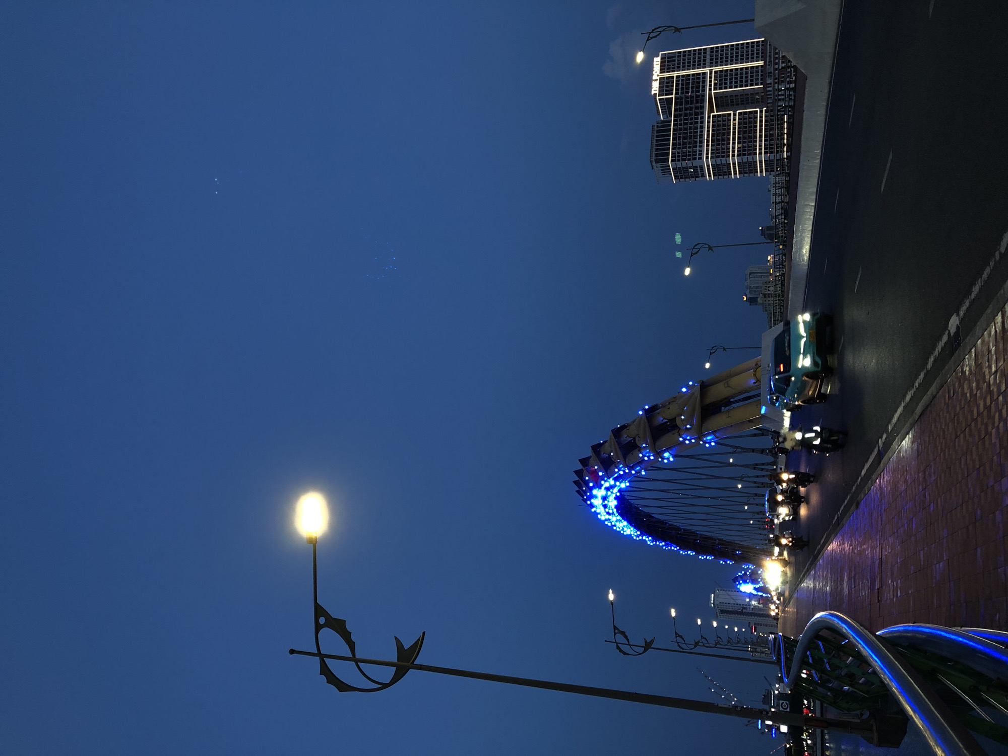 The image captures the Dragon Bridge in Da Nang, Vietnam at dusk or night. The iconic dragon-shaped bridge is illuminated with vibrant blue lights, with its arched structure prominently featured. A streetlamp with a decorative fish-shaped post stands to the left of the bridge, casting a warm, yellow glow. The bridge is open to traffic, with several motorcycles and a car visible on the roadway. The sky is a deep, clear blue, suggesting twilight or early night. In the background, modern buildings, including one labeled "THE PONTE," stand illuminated, adding to the urban cityscape. The perspective is from a slightly low angle, looking up towards the bridge and the sky. The foreground shows a paved walkway with a metal railing, also reflecting some of the blue light from the bridge.