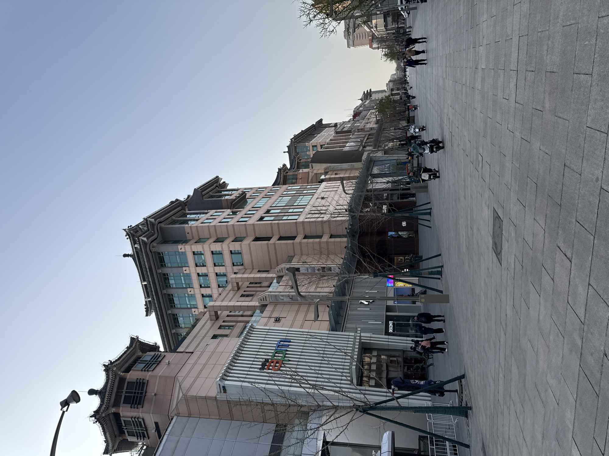 The image depicts a street scene in the Dongcheng District of China, characterized by a wide, gray-paved pedestrian area running along the right side of the frame. On the left, a series of multi-story buildings with traditional Chinese architectural elements, such as curved roofs and ornate details, line the street. Some of these buildings house commercial establishments, indicated by signs and storefronts.
People are visible both individually and in groups, walking along the paved area. Several individuals are on scooters. The sky is light blue. Sparse trees are planted along the street, with bare branches suggesting a winter or early spring season. Streetlights and other urban infrastructure are also present.
