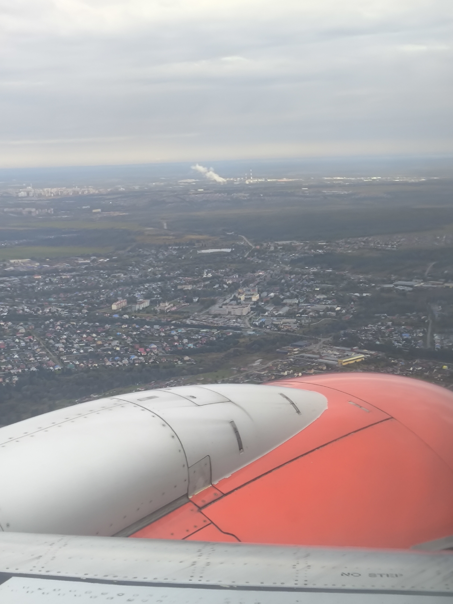 An aerial view captures a sprawling urban landscape under a cloudy sky, with the edge of an airplane's engine cowling in the foreground. The engine is predominantly white with a bright orange section, showing visible rivets and seams. Below the cowling, a grey wing surface with similar detailing is partially visible. The city below features a dense arrangement of buildings, including apartment blocks and numerous smaller houses with varied roof colors, indicating a residential area interspersed with some industrial or commercial structures. Trees and green spaces are scattered throughout the urban sprawl. In the distance, a plume of white smoke rises from what appears to be industrial facilities, suggesting the presence of a power plant or factory. The overall atmosphere is subdued due to the overcast conditions.