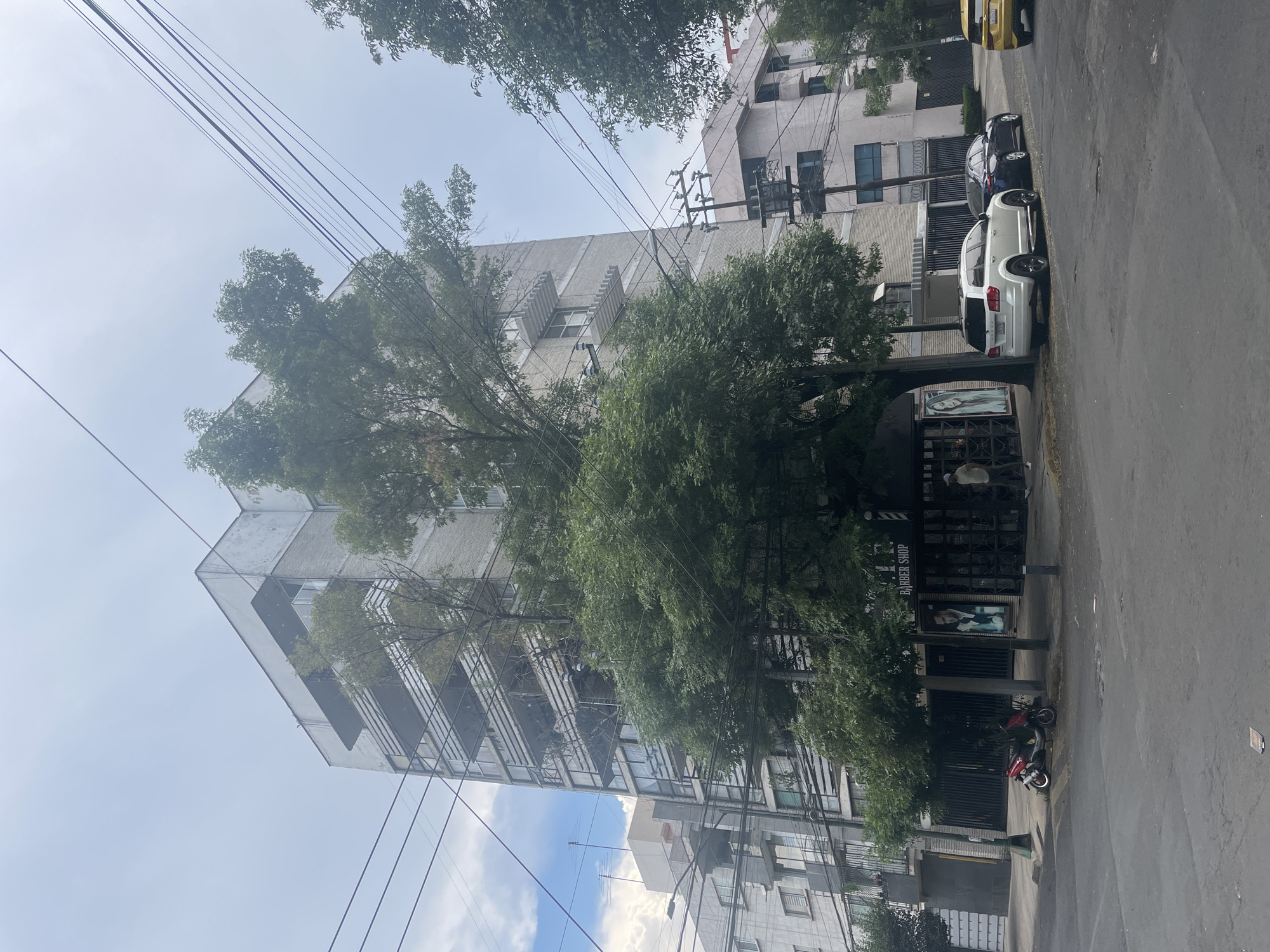 The image depicts a street scene in Mexico City, Mexico. The view is from street level, showing a tall, modern building with multiple balconies, partly obscured by a large tree. Overhead, a web of utility wires crisscrosses the sky.
Parked along the right side of the street are several cars: a white SUV, a dark-colored sedan, and the rear end of a yellow vehicle. Further down the street, there is a barbershop with a visible sign and advertisements. A red and black scooter is parked in front of the barbershop.
The weather appears overcast with a partly cloudy sky visible in the background. The architecture suggests a contemporary urban environment within Mexico City.