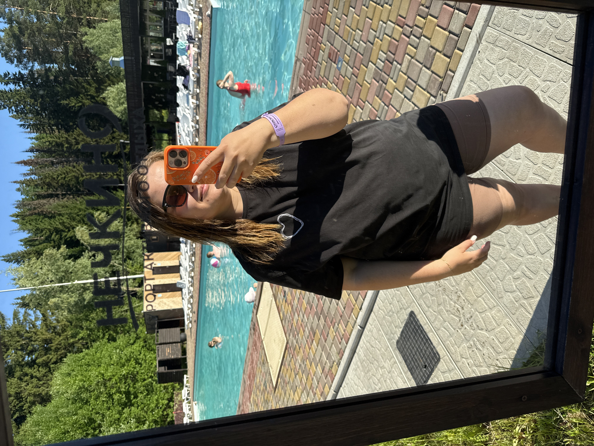 A woman in a black t-shirt and brown shorts takes a mirror selfie by a swimming pool. She is wearing sunglasses and holding an orange smartphone. In the background, people swim in the pool and sunbathe on lounge chairs. The location is identified as Nechkino, Russia, by a sign in the background that reads "Нечкино" and "ПОРОГ АКТИВ". The setting appears to be a resort or recreational area with trees and a clear sky.