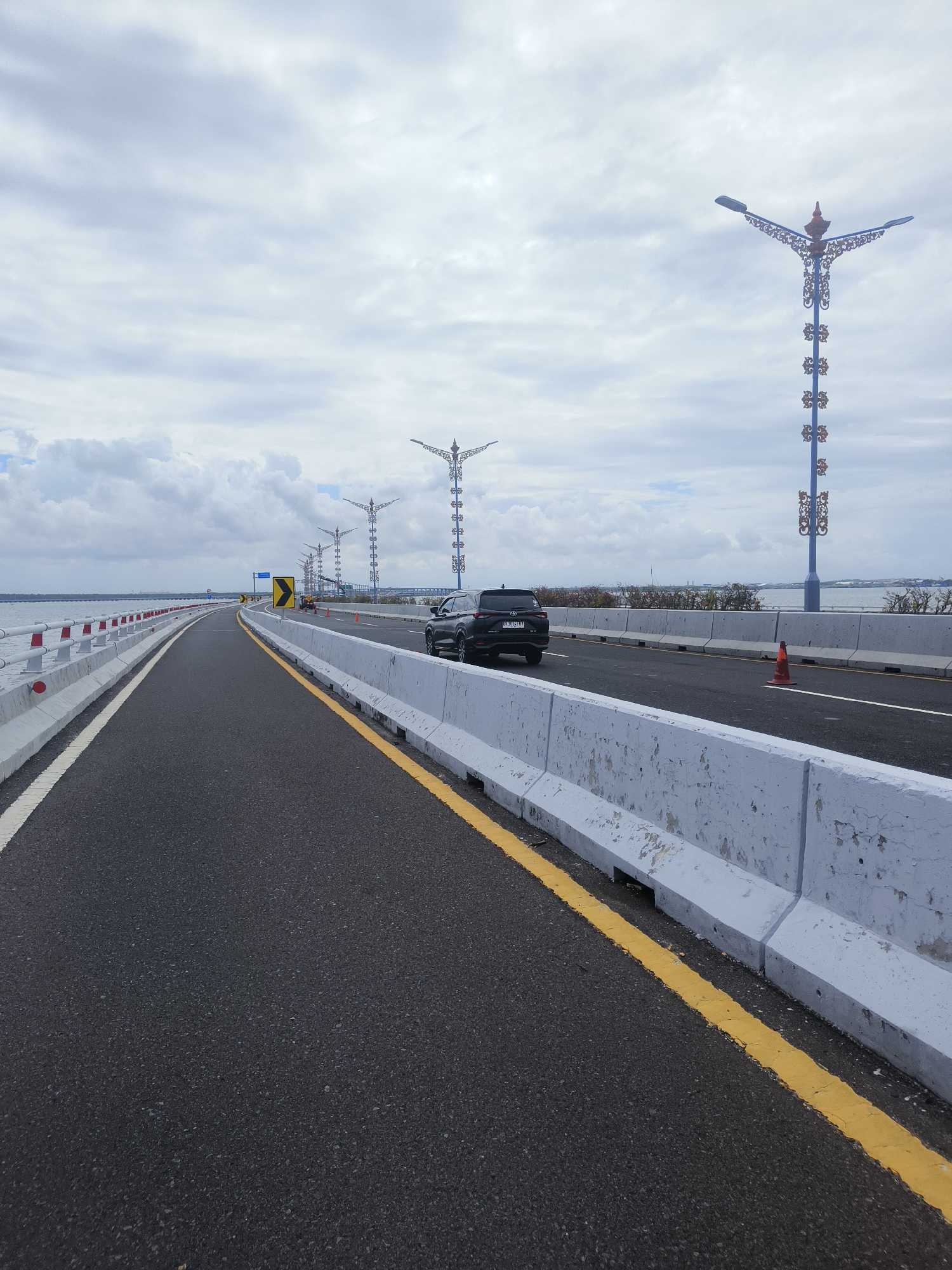 The image depicts a perspective view of a modern bridge, likely the Mandara Toll Road, in Kuta Selatan, Indonesia. The road surface is asphalt, clearly marked with white and yellow lines. On the left side, a white barrier with red and white reflective markers separates the road from a body of water. On the right, a continuous concrete barrier lines the road.
Several ornate streetlights with intricate blue designs and red accents are visible, spaced along the right side of the bridge. A black SUV is in motion on the right lane of the bridge, its rear visible to the viewer. Further down the bridge, more streetlights and a distant cityscape can be observed, partially obscured by the overcast sky. The weather appears to be cloudy, casting a soft light over the scene. The overall impression is one of contemporary infrastructure and a tranquil, expansive environment.