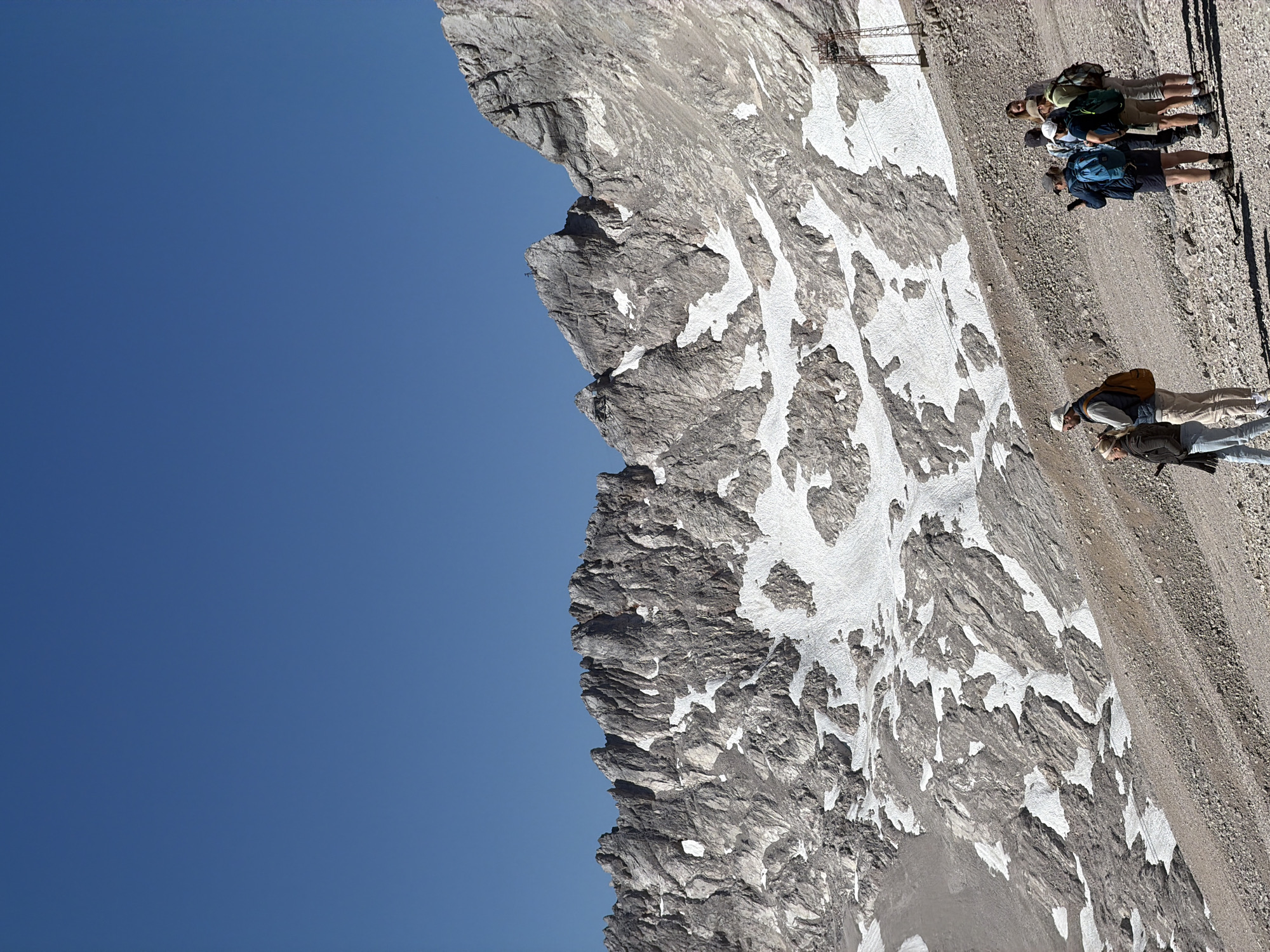 The image shows a group of people on a rocky, gravelly slope with patches of snow. In the background, there is a rugged mountain range under a clear blue sky. A cable car pylon is visible on the right side of the image. The people are dressed in outdoor clothing and appear to be hikers or tourists. Some are looking at their phones, while others are engaged in conversation. The scene is set in a mountainous, alpine environment, likely at a high elevation.