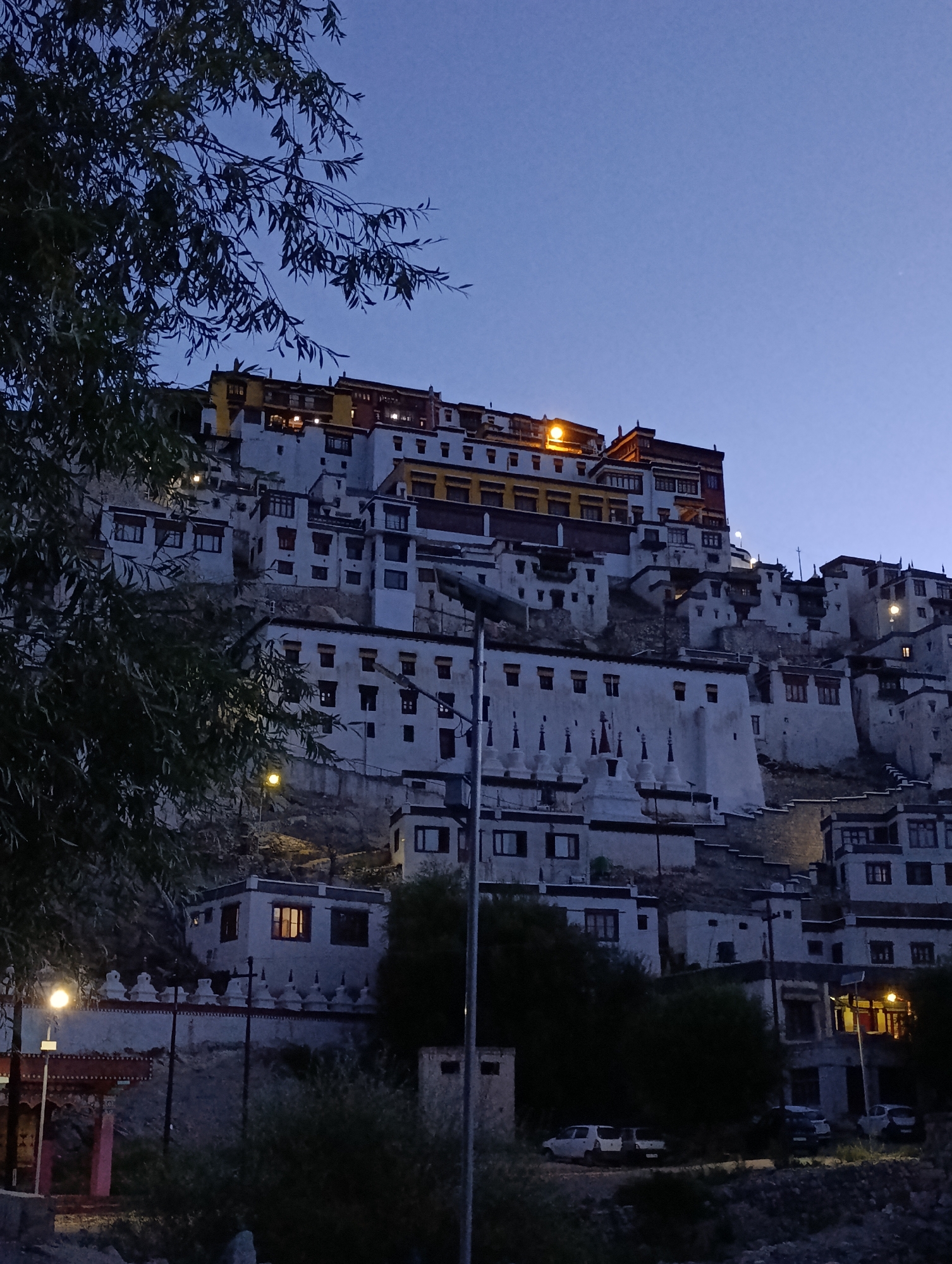 The image depicts the Thiksey Monastery in Ladakh, India, during twilight. The monastery, a multi-story complex, is built on a hillside and characterized by its white-washed buildings with red-trimmed windows and yellow accents on the upper levels. Several small stupas are visible at the lower sections of the complex. The sky is a deep blue, indicating the transition from day to night. Trees with dark green foliage are in the foreground, partially obscuring the view of the monastery. Artificial lighting, visible as warm yellow glows from windows and street lamps, illuminates parts of the structure and the surrounding area. In the lower right portion of the image, several parked cars are visible, suggesting the presence of modern amenities and visitors or residents. The overall scene conveys a sense of serenity and the grandeur of the monastic architecture against the darkening sky.