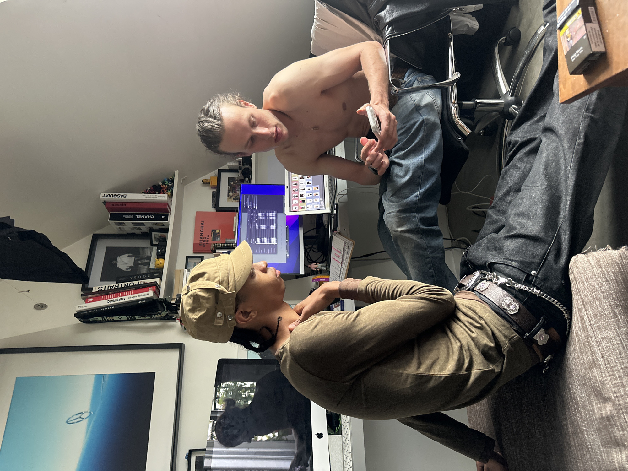 The media file captures an indoor scene, likely within an apartment or studio in Paris, France, during daylight hours. Two individuals are engaged in a casual interaction.
On the left, a shirtless man with light-colored hair is seated on a black office chair, positioned at a desk with two computer monitors, including an Apple iMac. He wears blue jeans and holds a clear, small object, possibly a vape, in his hand, looking down towards the other person.
On the right, another individual with darker skin and hair, wearing a long-sleeved olive-toned top and a matching cap, is seated on the armrest of a light-colored sofa. This person's left hand is resting on their neck, and they are looking up towards the seated man. They wear dark trousers with a distinctive belt featuring silver buckles, chains, and decorative embellishments.
The background includes a bookshelf filled with books, with visible titles such as "BASQUIAT," "CHANEL," "David Bailey," "SHANGHAI WING SHYA," "PORTRAITS," and "BOOBA." Framed artworks or photographs adorn the walls. A pack of "PHILIP MORRIS" cigarettes is visible on a wooden surface. The room features an angled ceiling, indicative of an attic or mansard-style space. The overall atmosphere suggests a relaxed, personal setting with the two individuals engaged in a focused exchange.